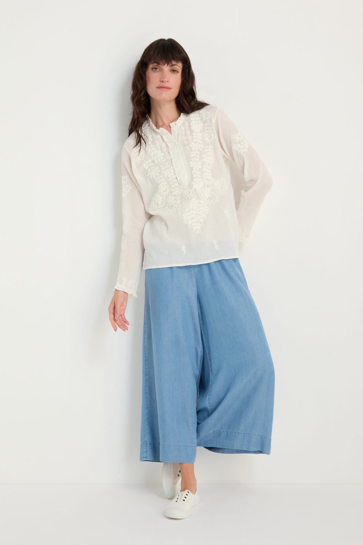 A woman stands against a white wall wearing the Embroidered Cotton Top in natural white, paired with wide-leg light blue pants and white sneakers.