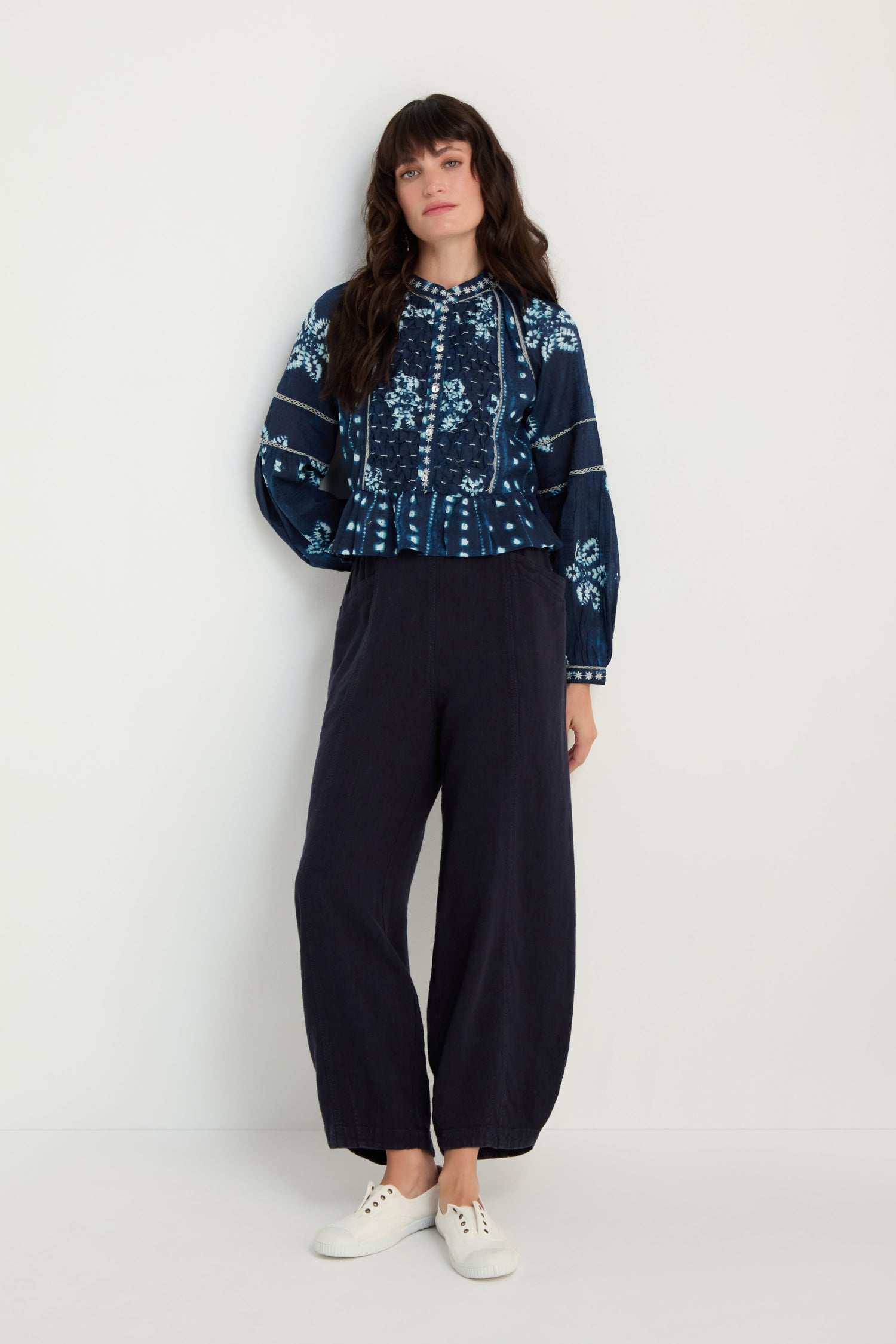 A woman stands against a white wall wearing the Mixed Shibori Shirt, a navy floral pure cotton long-sleeve blouse with embroidered trim, paired with wide-leg dark trousers and white sneakers.