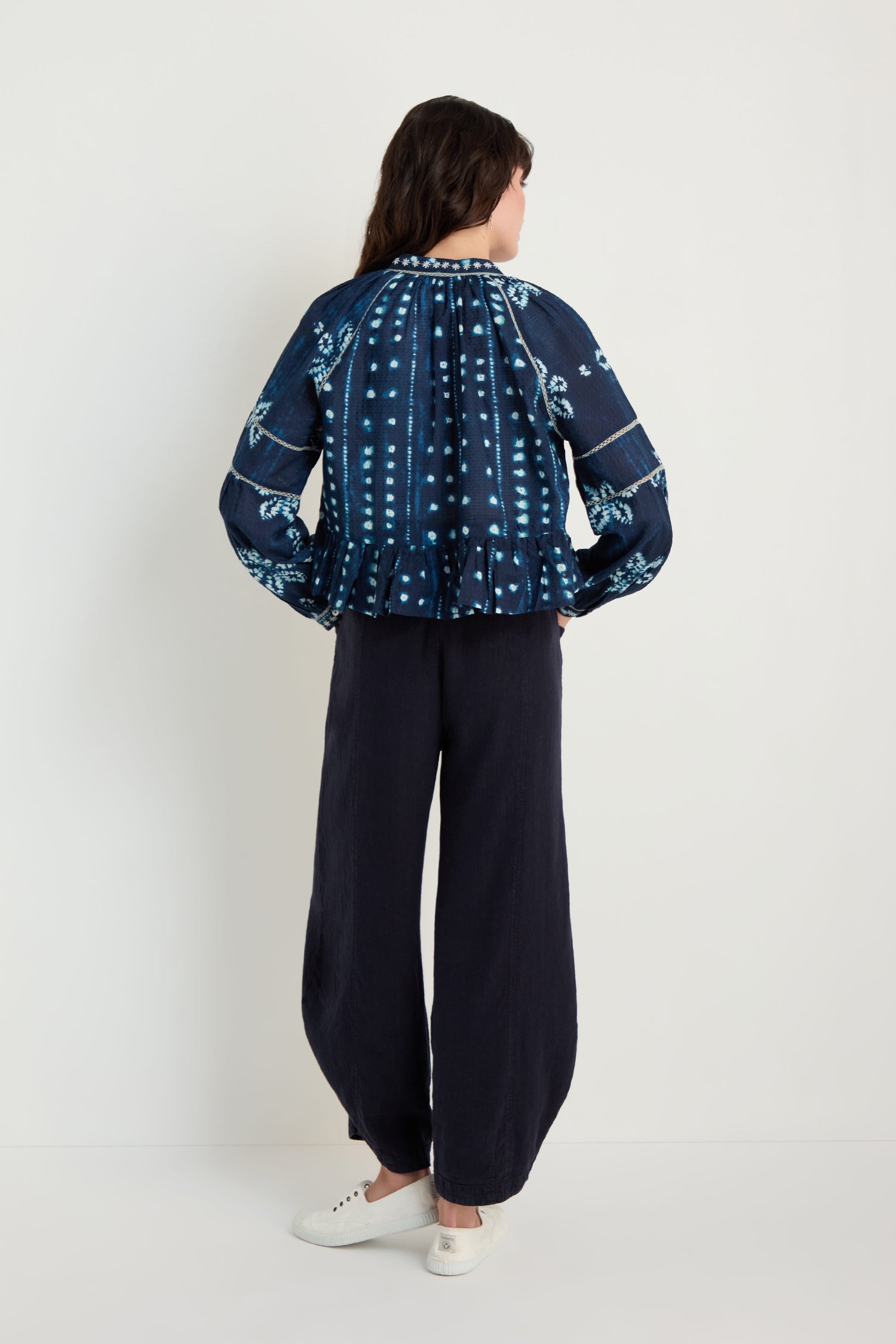 A woman stands against a white wall, facing away, wearing the Mixed Shibori Shirt in dark blue with embroidered trim, paired with loose black pants and white slip-on shoes.