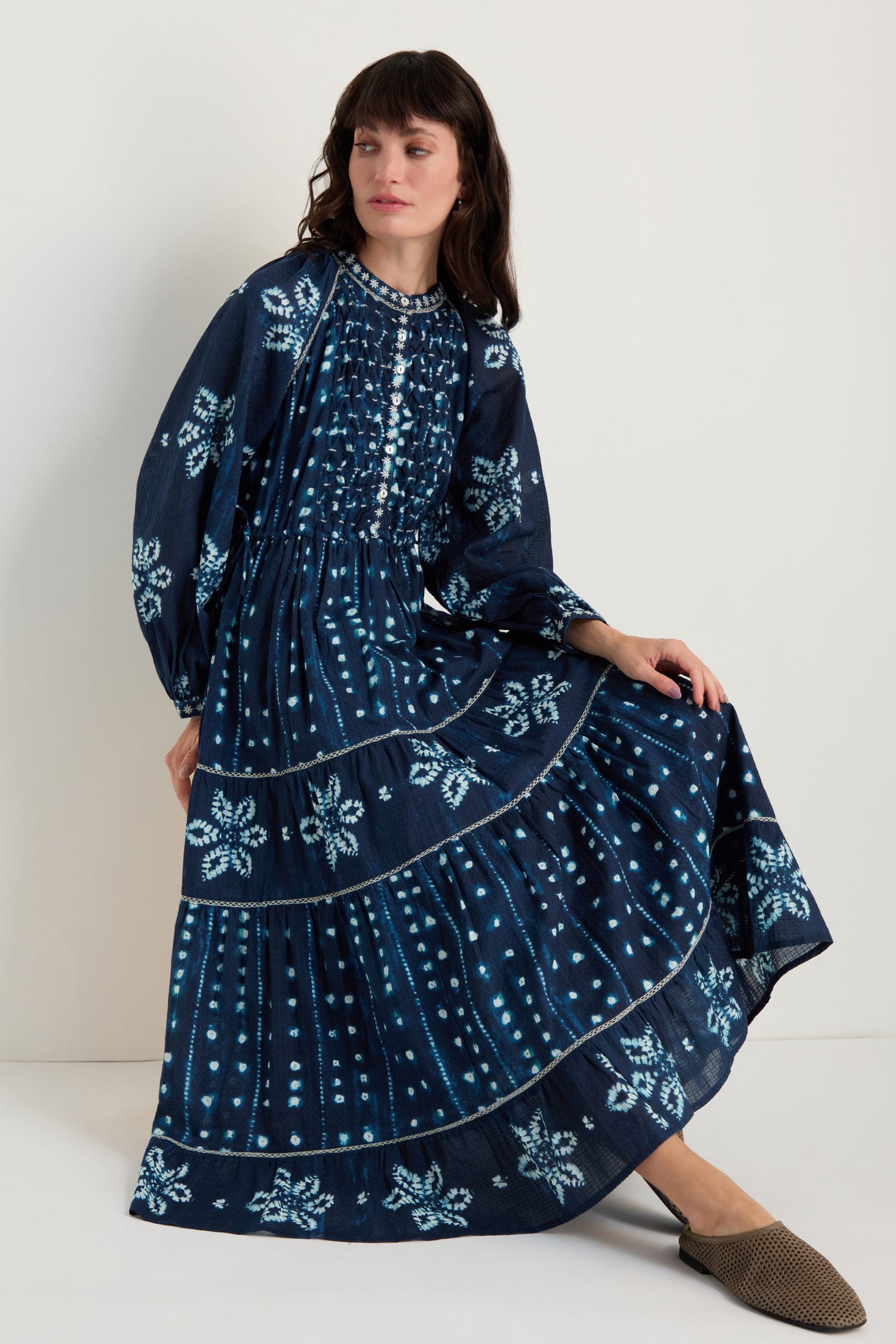 A woman sits against a white wall wearing the Mixed Shibori Dress—a long-sleeve, navy blue pure cotton midi with light blue floral and dot patterns—and brown flats.