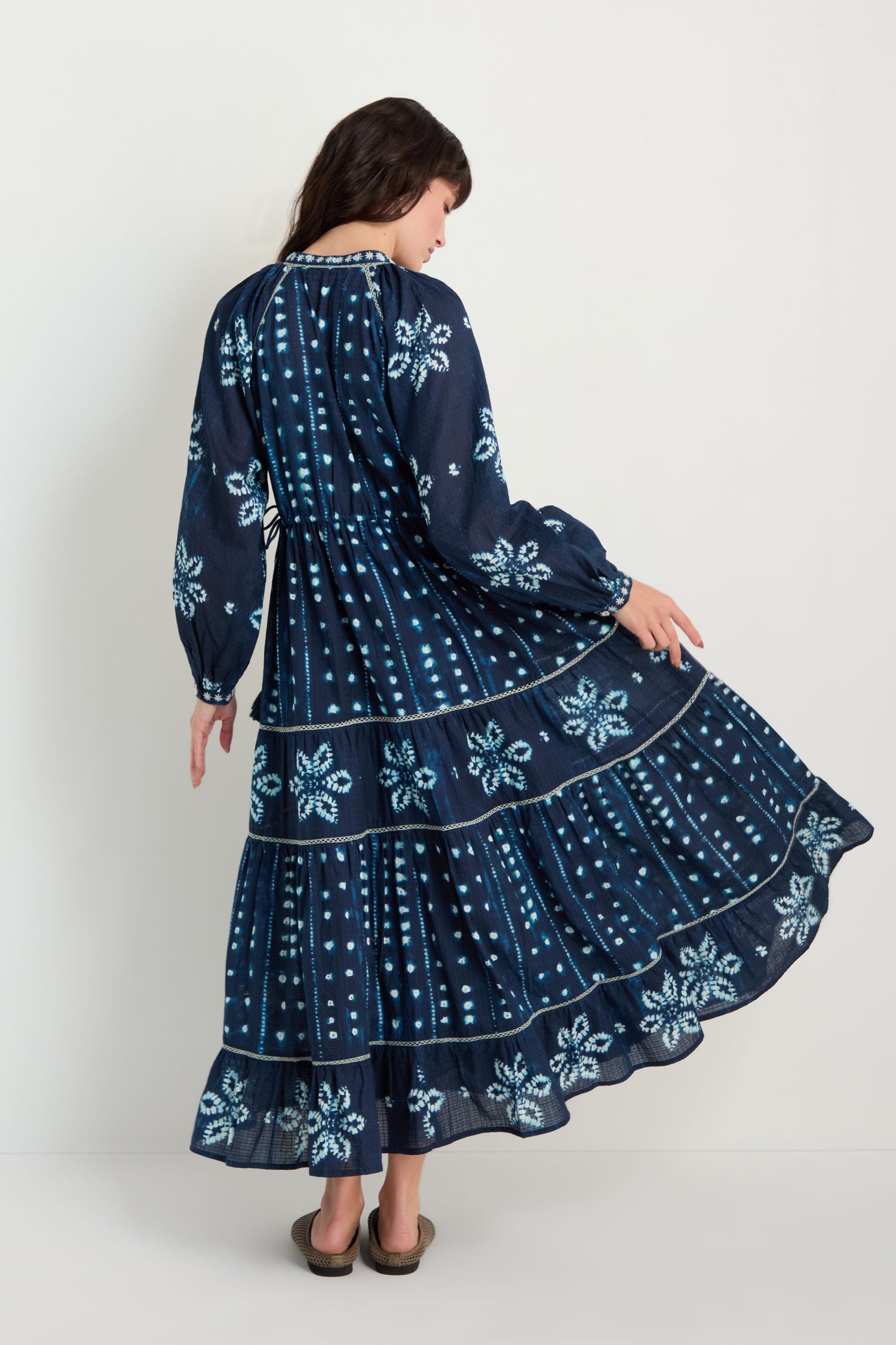 A woman stands facing away, holding out one side of the skirt while wearing the Mixed Shibori Dress—a long-sleeve, pure cotton navy blue maxi dress featuring white floral and dotted patterns.