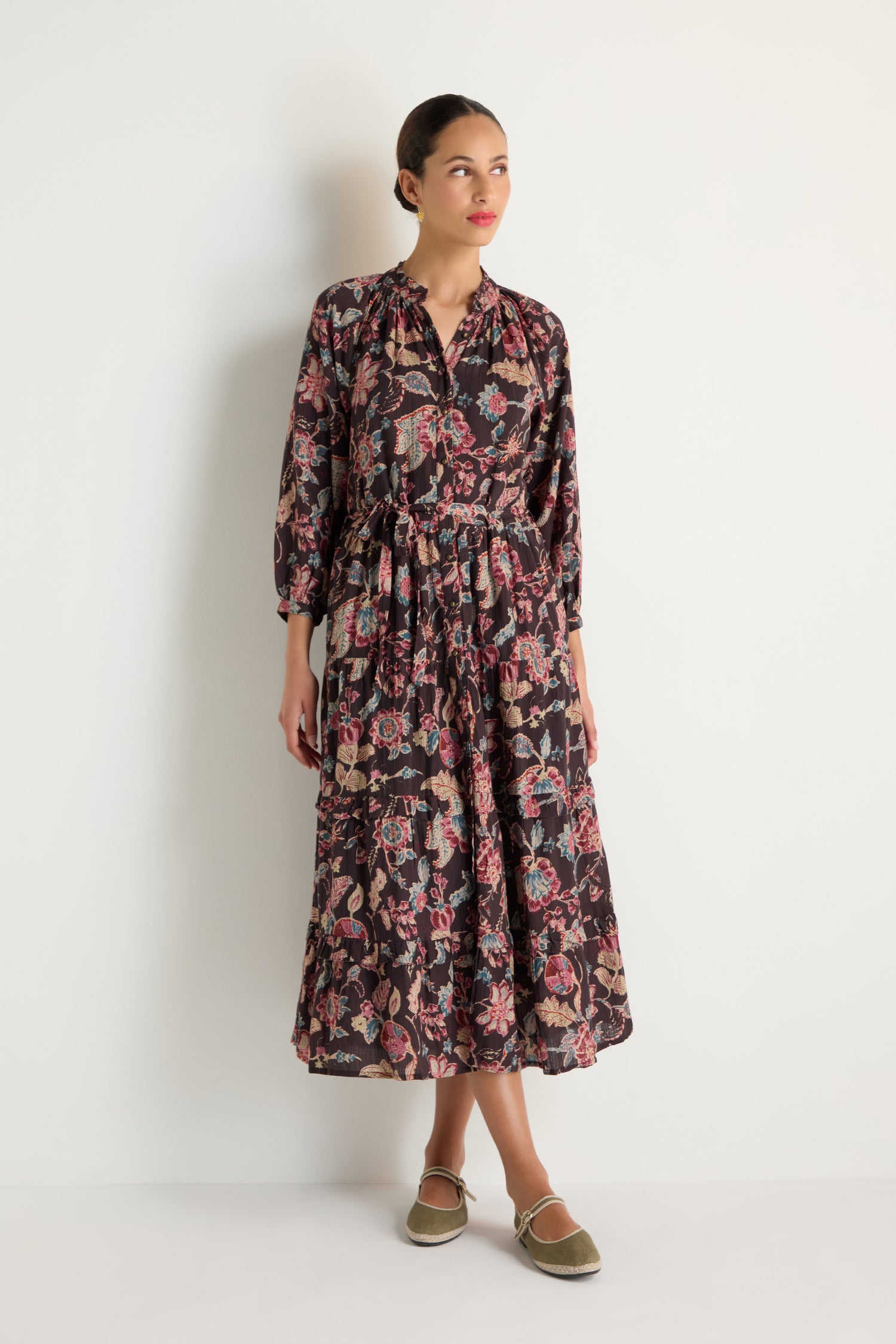 A woman stands against a white wall wearing the Iris Print Tiered Dress, an ankle-length, long-sleeved floral and paisley dress with a gathered waist, paired with brown espadrille flats.