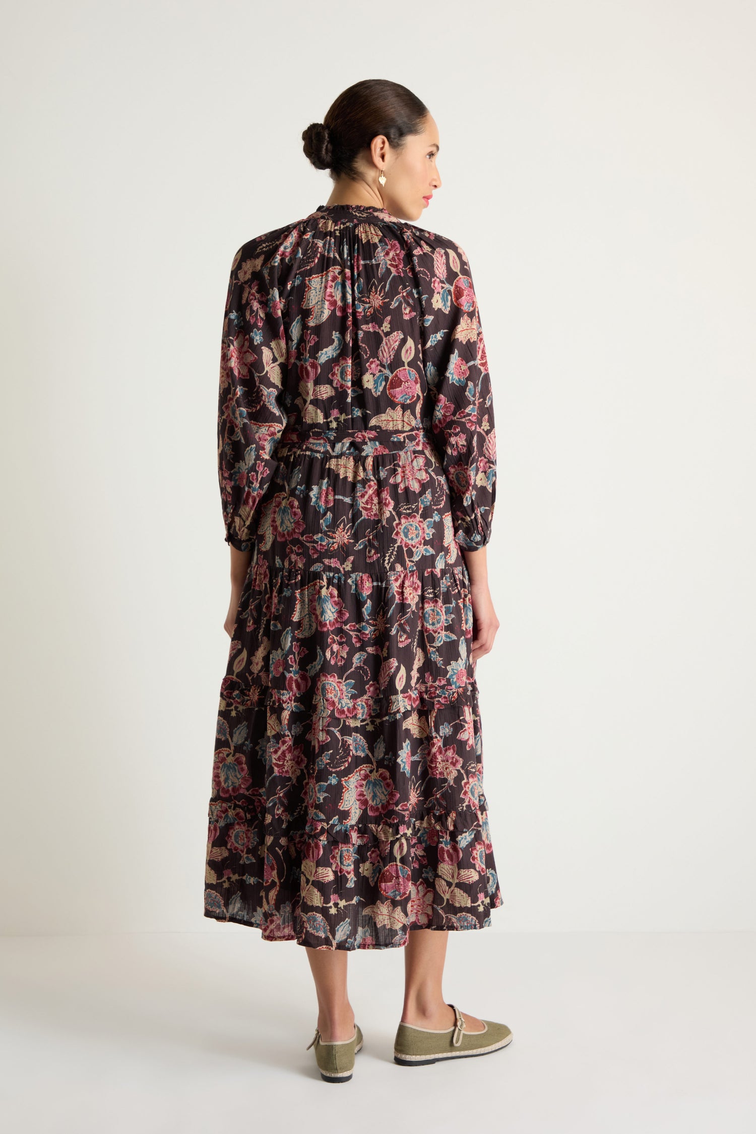 A woman with her hair in a bun stands against a plain background, facing away, wearing the long-sleeved Iris Print Tiered Dress with floral and paisley patterns and slip-on shoes.