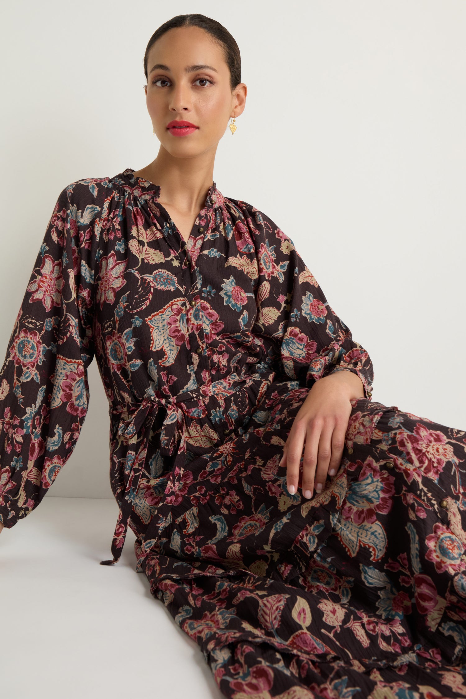 Woman sits against a plain white background wearing the Iris Print Tiered Dress, a long-sleeved dress featuring floral and paisley prints with a tie waist.
