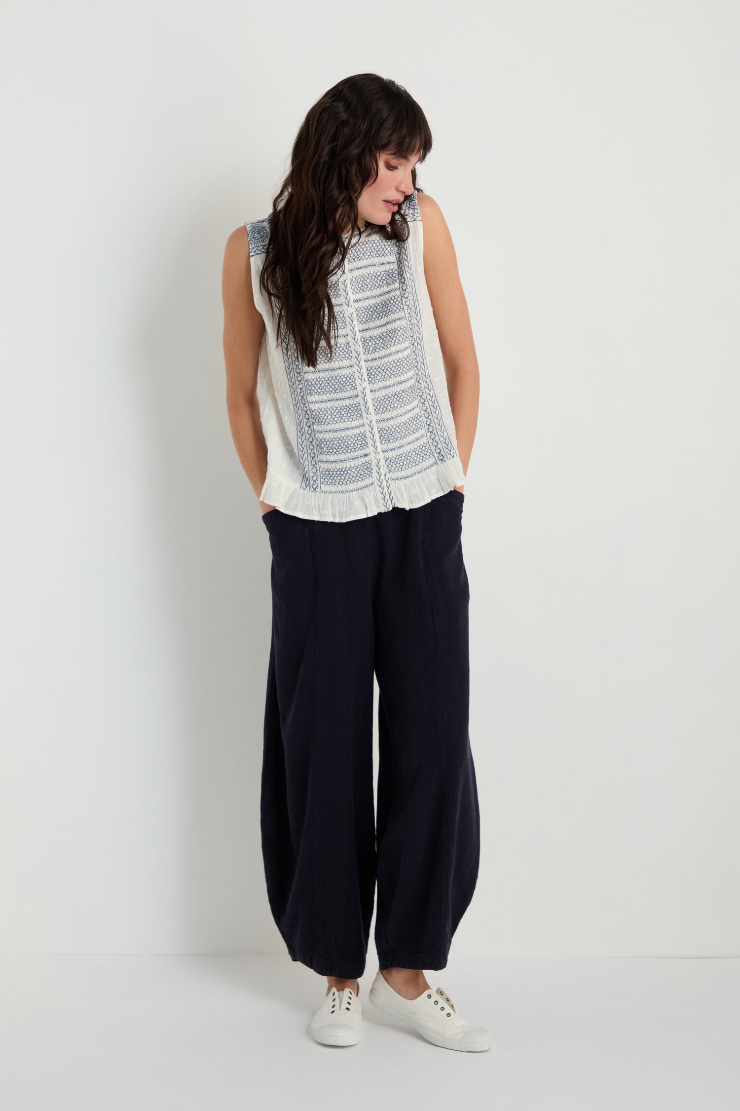 A woman stands against a plain white wall, wearing the Embroidered Button-Through Top, loose dark trousers, and white sneakers, gazing down and slightly to the side.