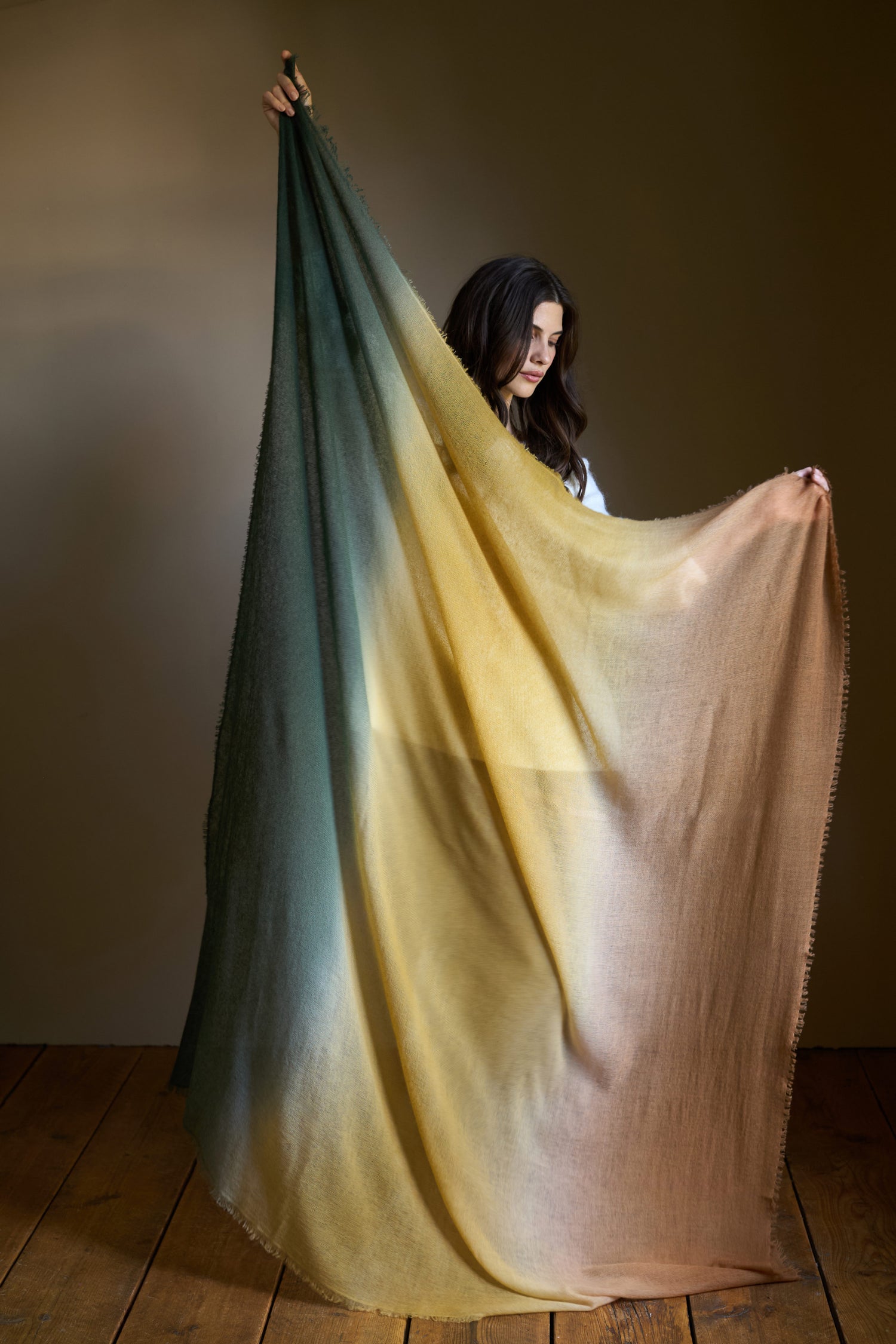 A woman stands on a wooden floor holding up the Soft Colourblend Cashmere Shawl, a large, sheer luxury wrap with green, yellow, and brown gradient tones.