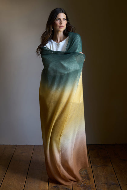 A woman stands on a wooden floor, wrapped in the Soft Colourblend Cashmere Shawl that fades from green to yellow to brown, with a white top underneath.