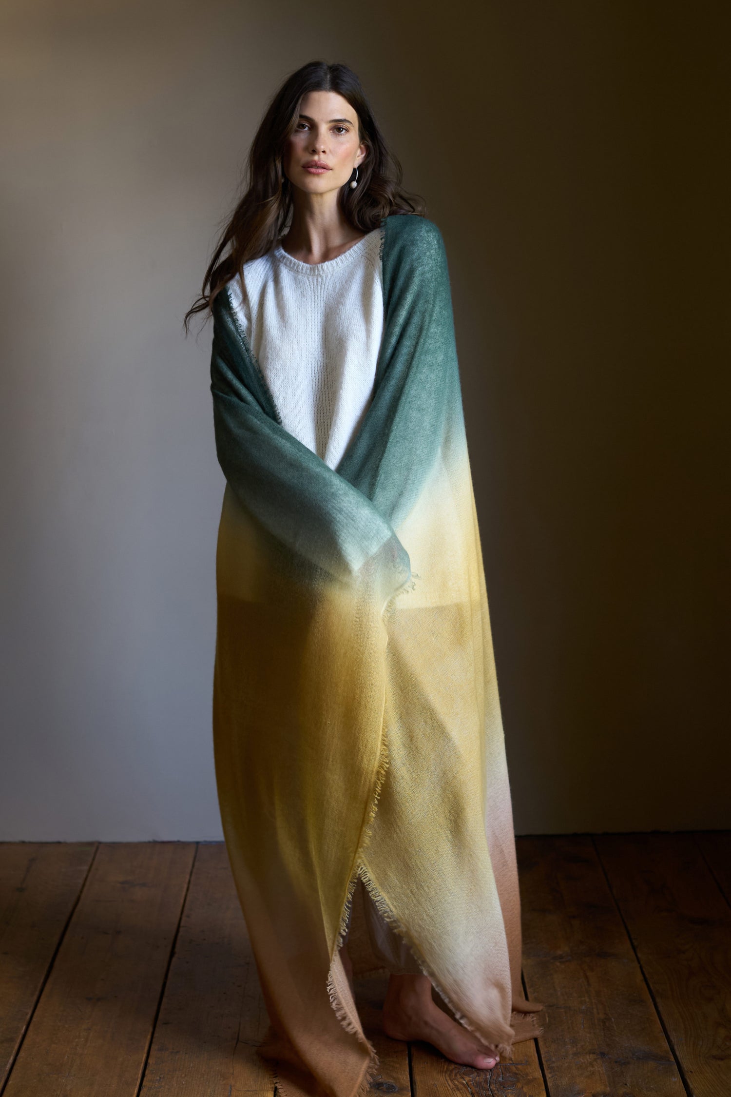 A woman stands on a wooden floor, wrapped in a Soft Colourblend Cashmere Shawl featuring green, yellow, and brown tones, wearing a white dress and gazing at the camera.