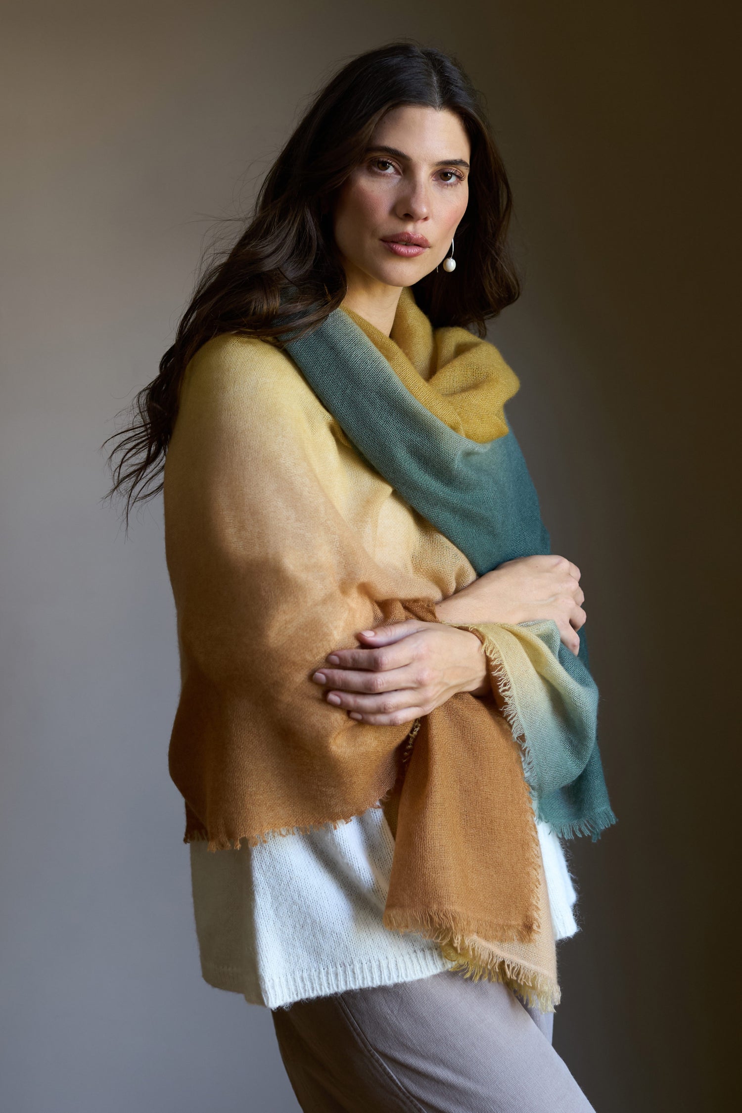 A woman with long dark hair wears a Soft Colourblend Cashmere Shawl in yellow, brown, and teal over a white top and beige pants, standing against a plain background.