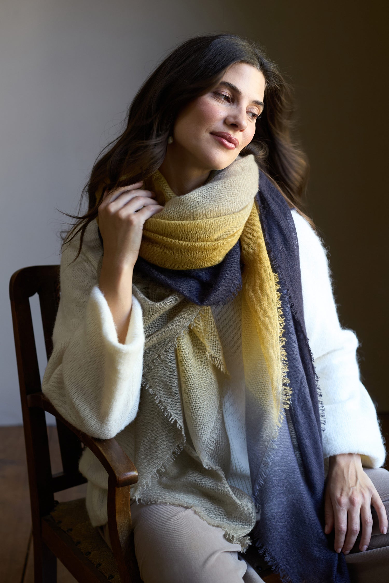 A woman with long brown hair sits on a wooden chair, wearing a white sweater and the Soft Colourblend Cashmere Shawl—an elegant Nepal-woven pure cashmere scarf in yellow, beige, and navy gradient tones.