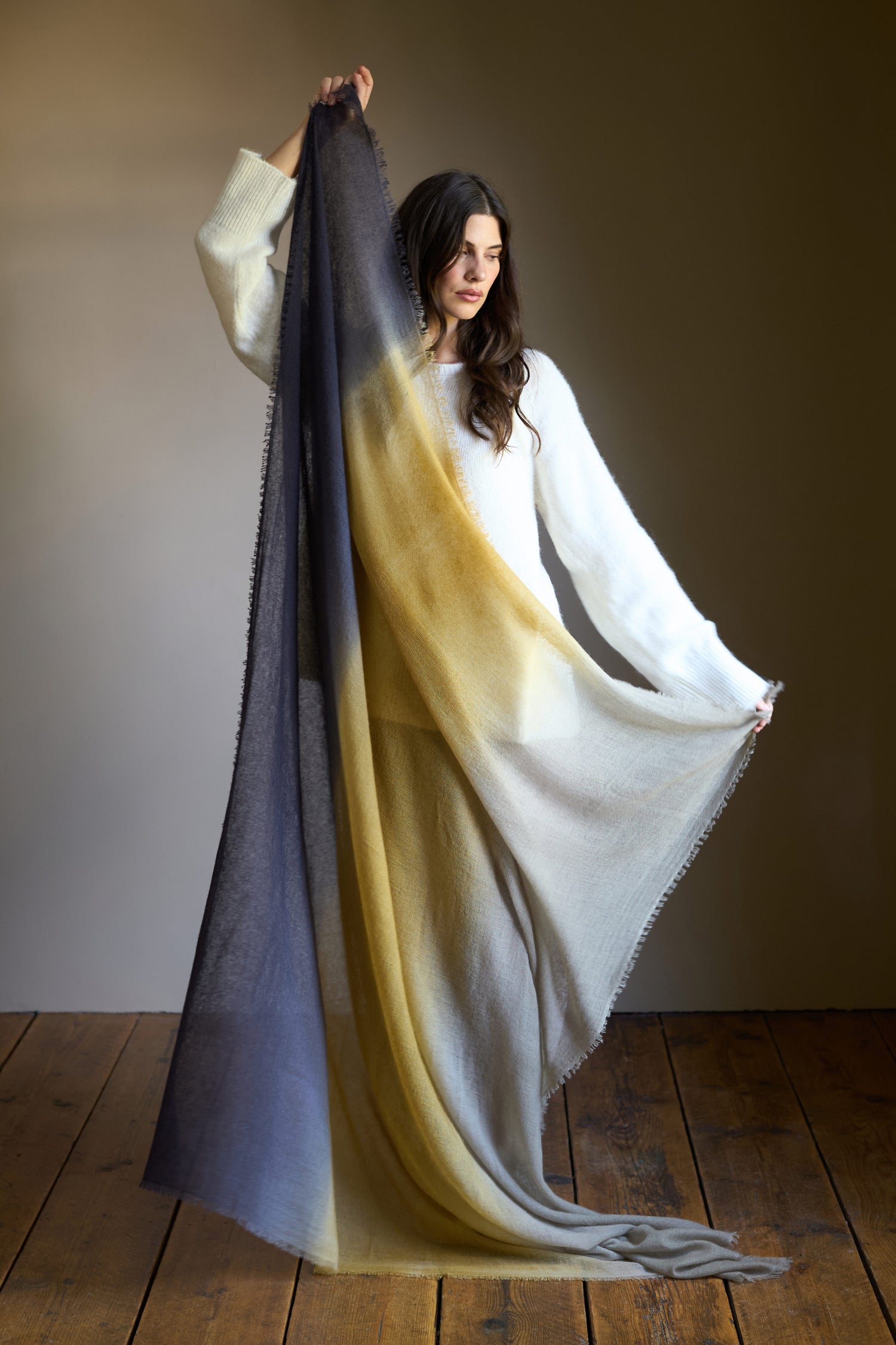 A woman in a white sweater stands on a wooden floor, displaying the Soft Colourblend Cashmere Shawl in ombré gray, yellow, and cream shades, highlighting its luxurious softness and exquisite Nepalese craftsmanship.