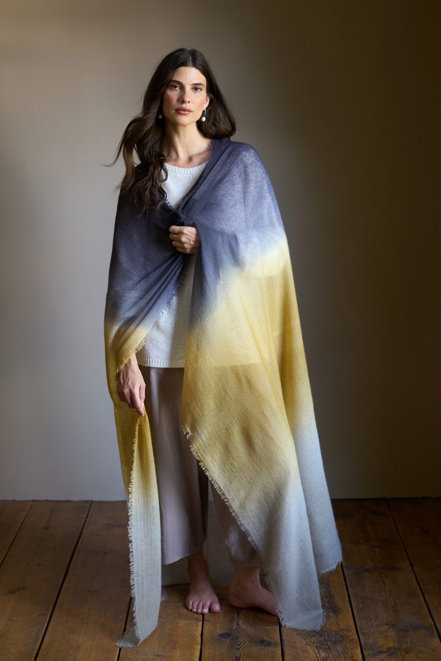 A woman stands barefoot on a wooden floor, wearing white and draped in the Soft Colourblend Cashmere Shawl, which features gradient shades of gray, yellow, and cream.