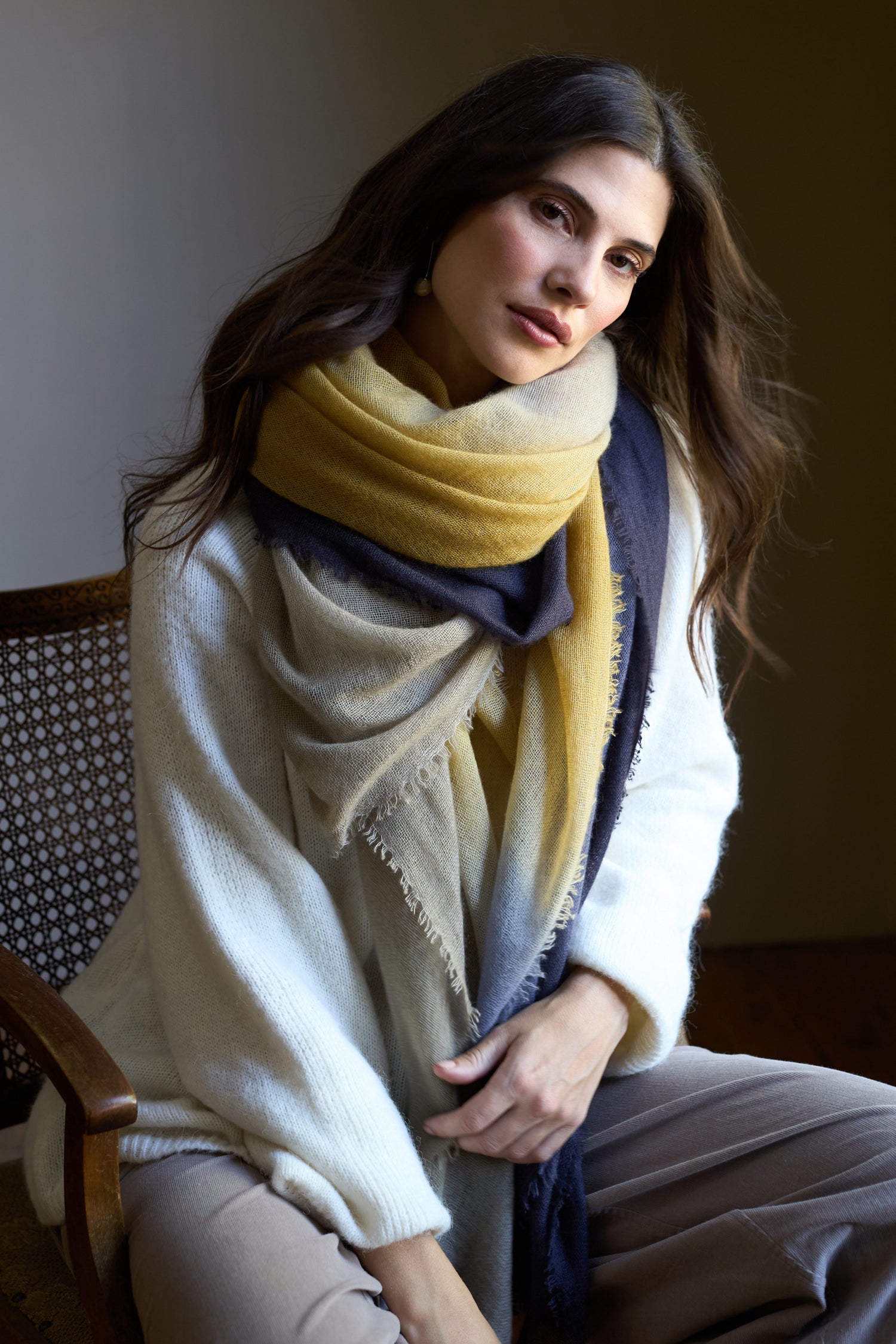 A woman sits on a chair wearing a cream sweater, beige pants, and a Soft Colourblend Cashmere Shawl in yellow and navy, looking slightly to the side.