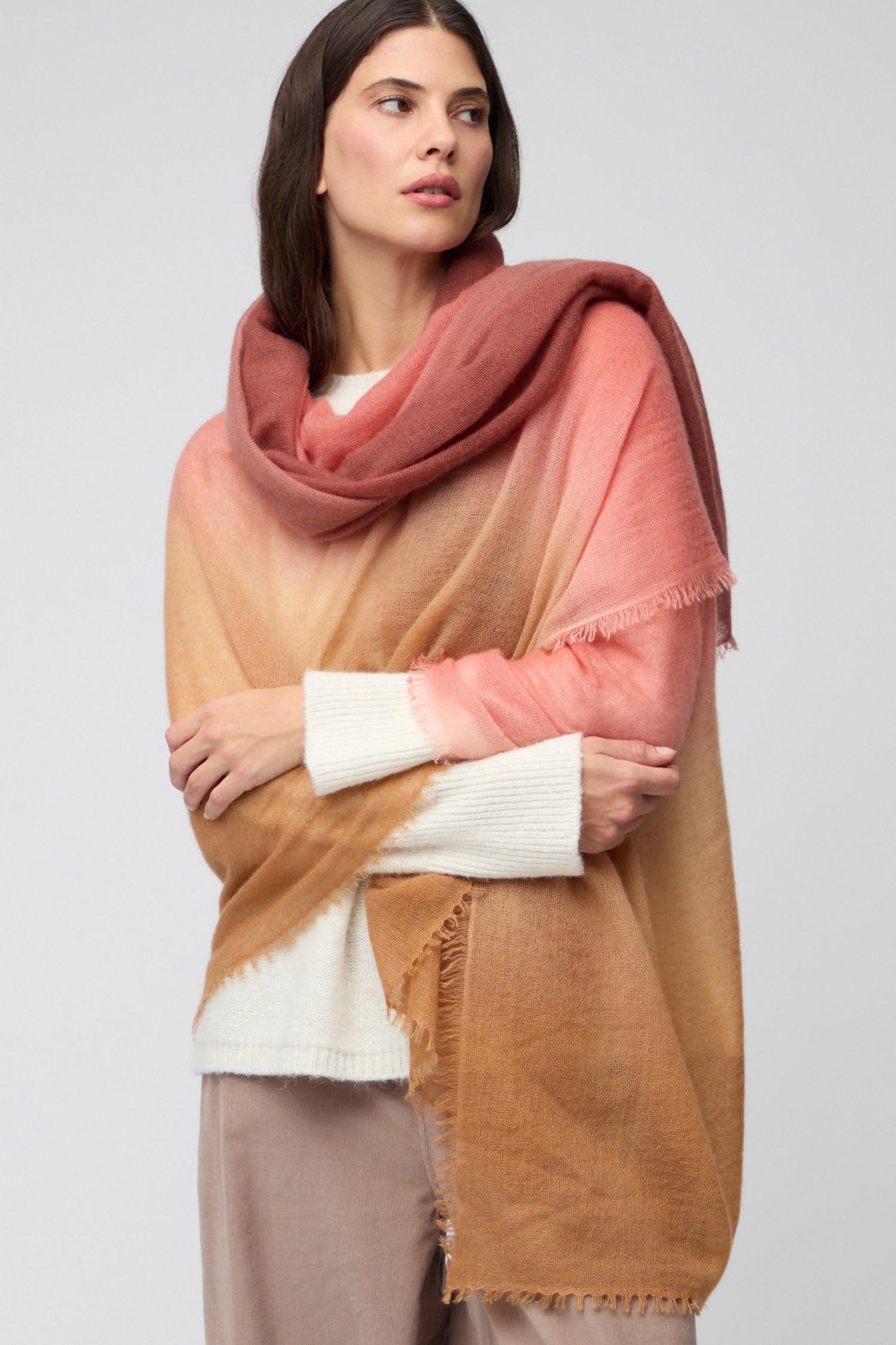 A woman wears the Soft Colourblend Cashmere Shawl, featuring a large ombré gradient in brown, tan, and rose over a white sweater and beige pants—a true luxury accessory for any wardrobe.