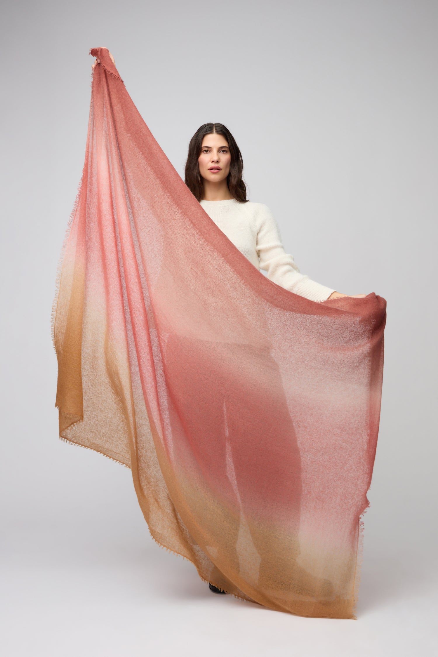 A woman in a cream sweater displays the Soft Colourblend Cashmere Shawl, a large semi-transparent accessory with an elegant ombré gradient in pink, beige, and tan—an exquisite touch of luxury against a plain background.