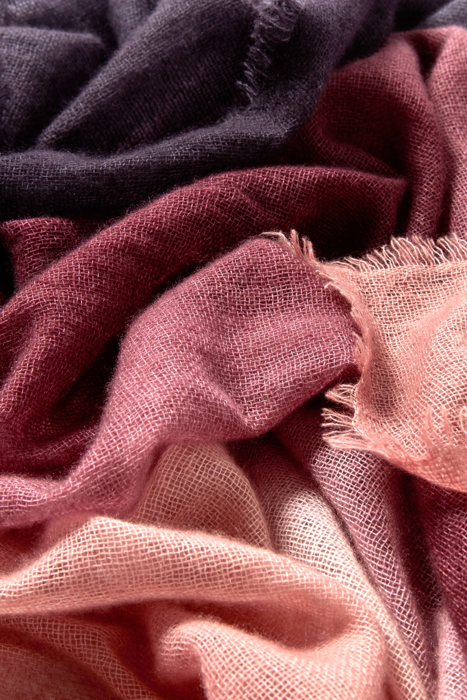 A close-up of the Soft Colourblend Cashmere Shawl highlights its layered, loosely woven fabric in deep purple, burgundy, mauve, and pale pink tones with textured, frayed edges—an elegant and timeless accessory.