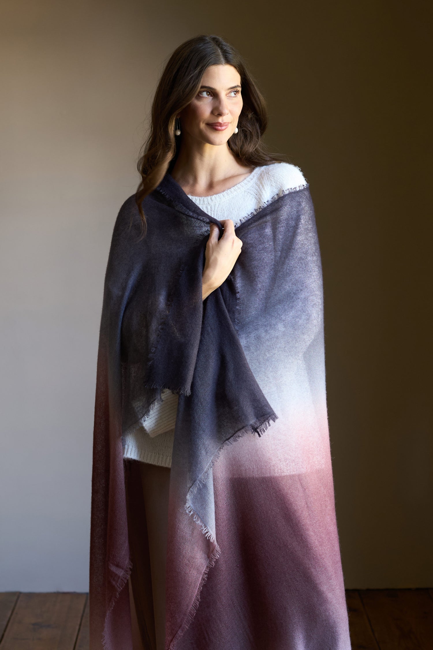 A woman with long brown hair in a white top wears the Soft Colourblend Cashmere Shawl—Nepal-woven pure cashmere in gradient gray and burgundy—while standing indoors against a plain background.
