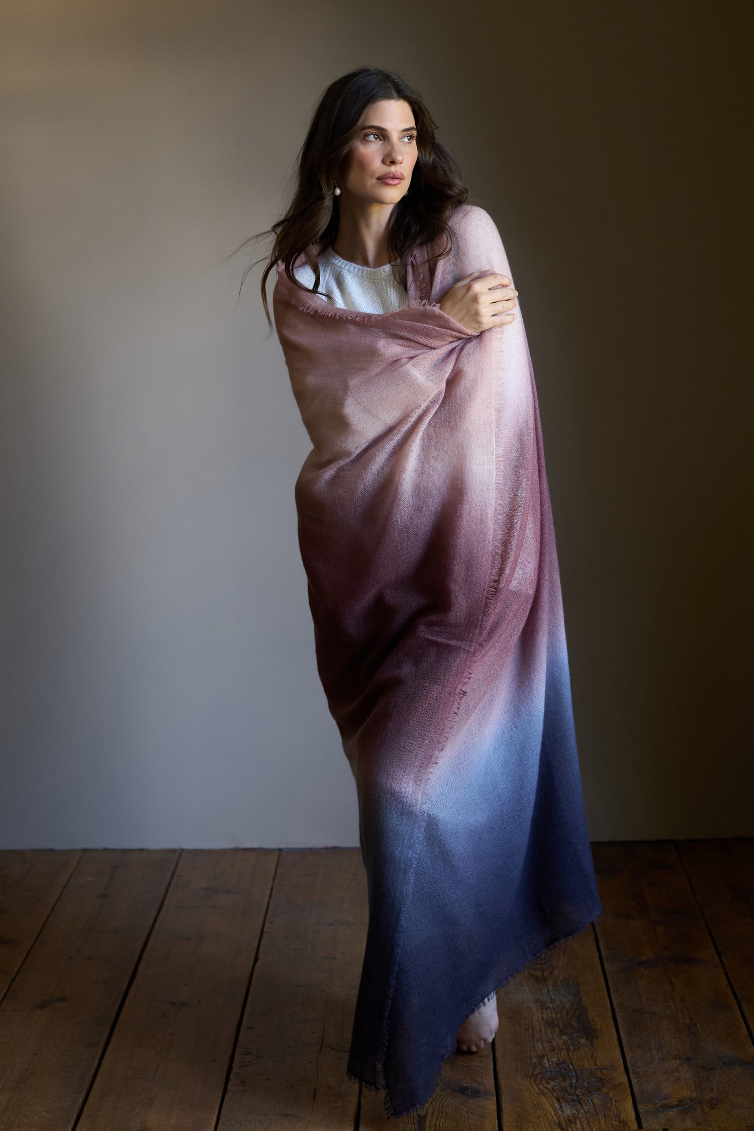 A woman stands on a wooden floor, draped in the Soft Colourblend Cashmere Shawl—a luxury wrap woven in Nepal from pure cashmere that features a pink-to-blue gradient—set against a neutral background.