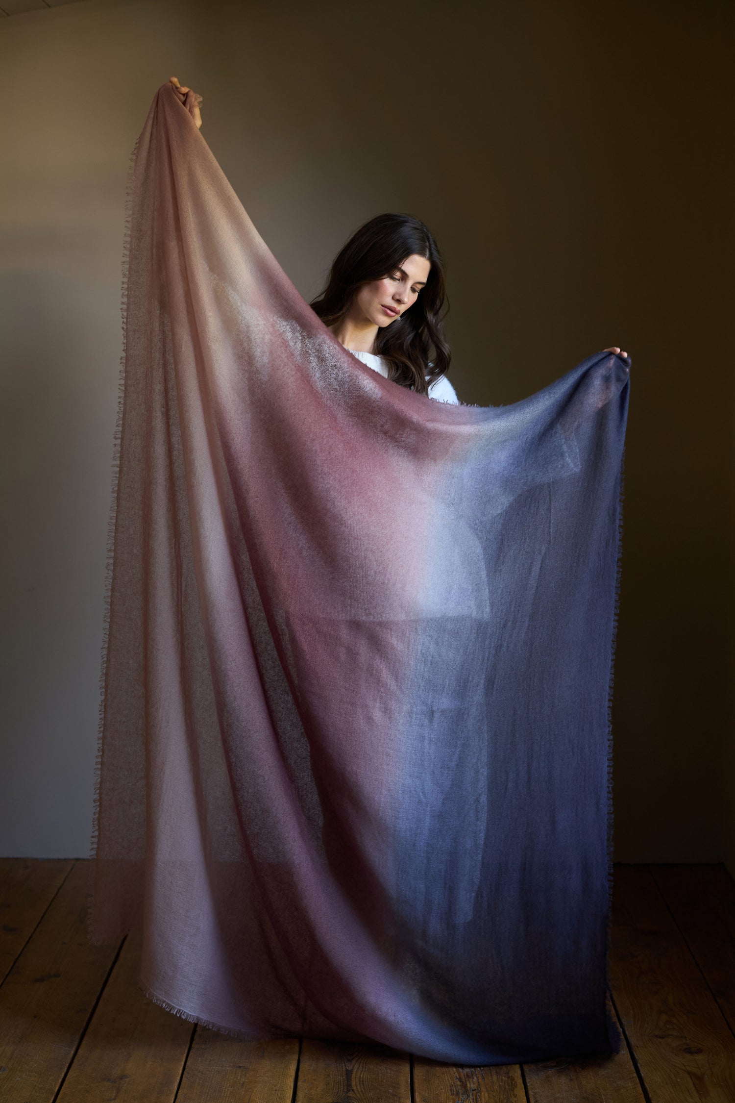 A woman stands on a wooden floor holding up the Soft Colourblend Cashmere Shawl, a large, sheer luxury wrap featuring a gradient from light brown to dark blue, designed with inspiration from traditional Nepalese weaving.