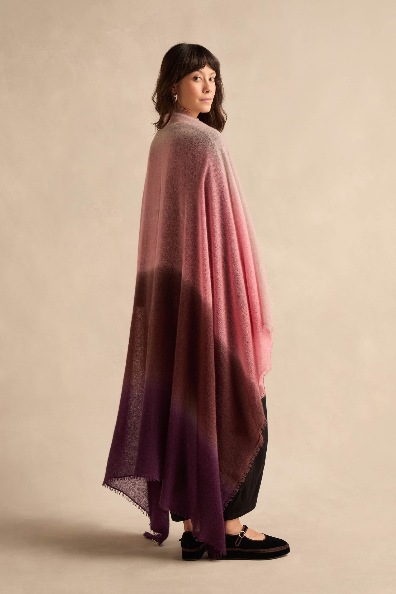 A woman in profile wears the Cherry and Pink Soft Colourblend Cashmere Shawl—woven in Nepal with pink, burgundy, and purple hues—draped over black pants and shoes, standing against a neutral background.