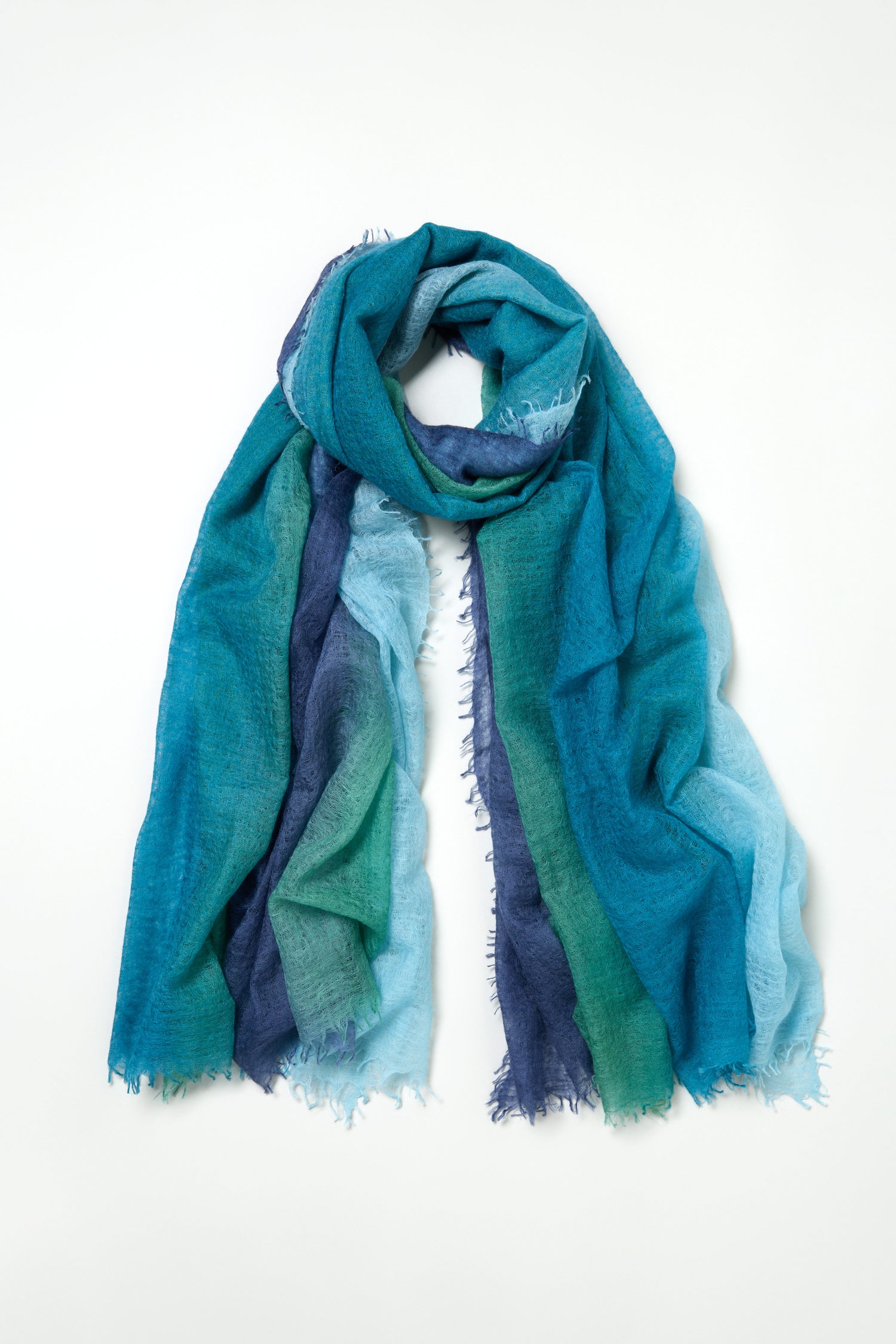 The St. Tropez Cashmere Shawl is a lightweight layering scarf with frayed edges, featuring blue and green hues that evoke breezy St Tropez style, shown laid flat on a white background.