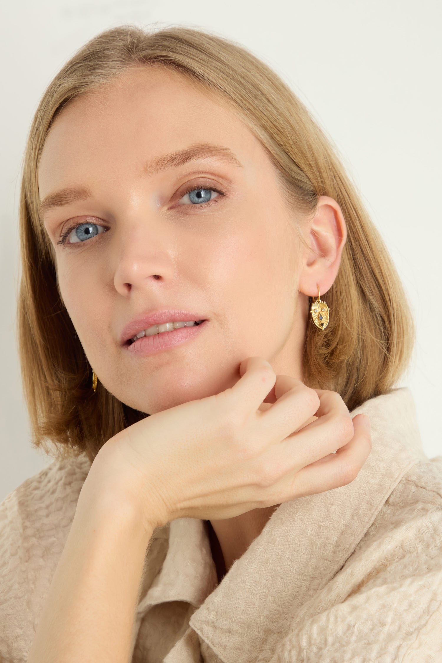 A woman with light skin and blonde hair, wearing a beige textured top, gazes at the camera with her chin resting on her hand. She accessorizes with Gold Plated Labradorite Mini Faces Earrings.