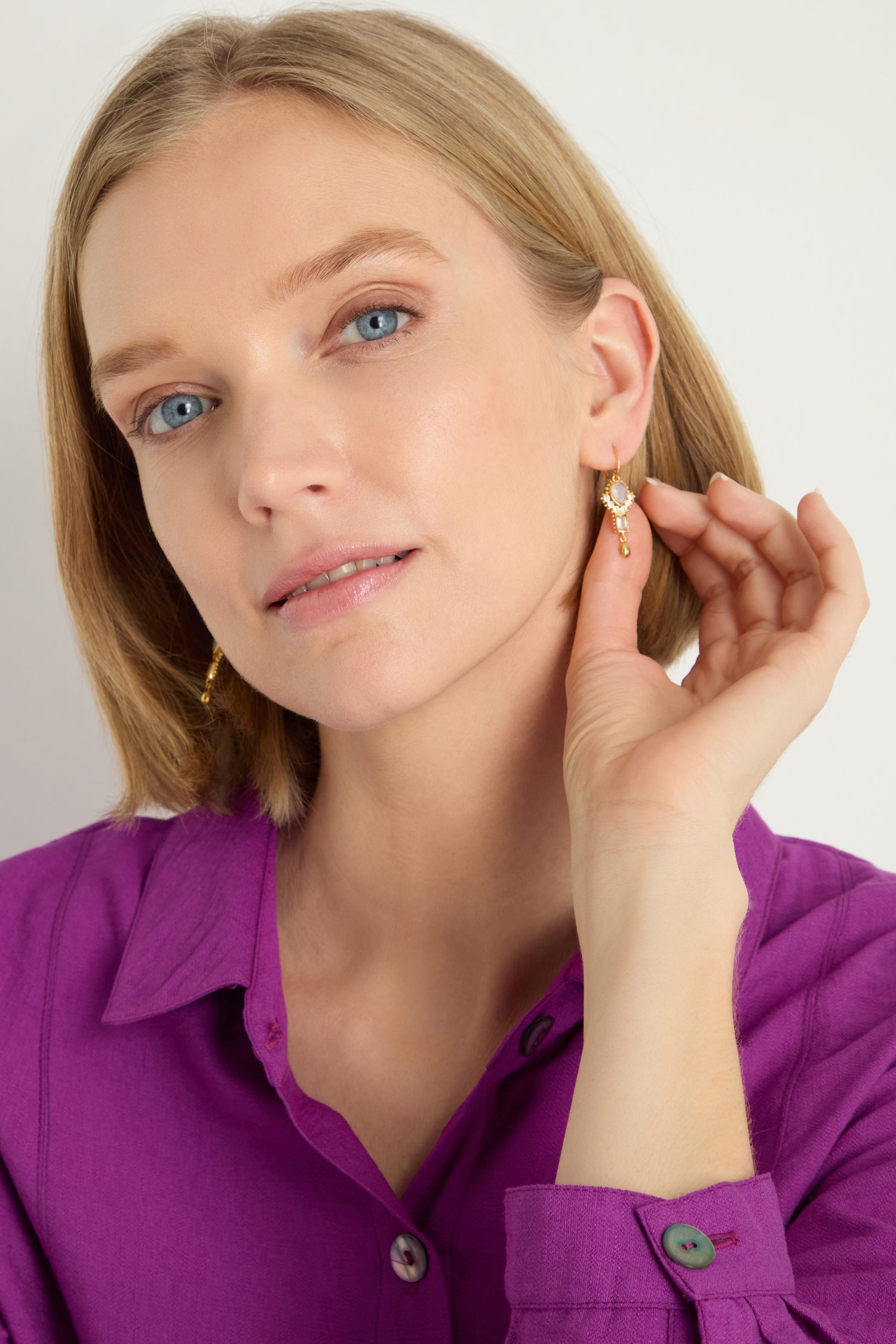 A woman in a purple shirt holds up the Gold Plated White Moonstone Ariel Earrings to her left ear, gazing confidently at the camera against a plain background.
