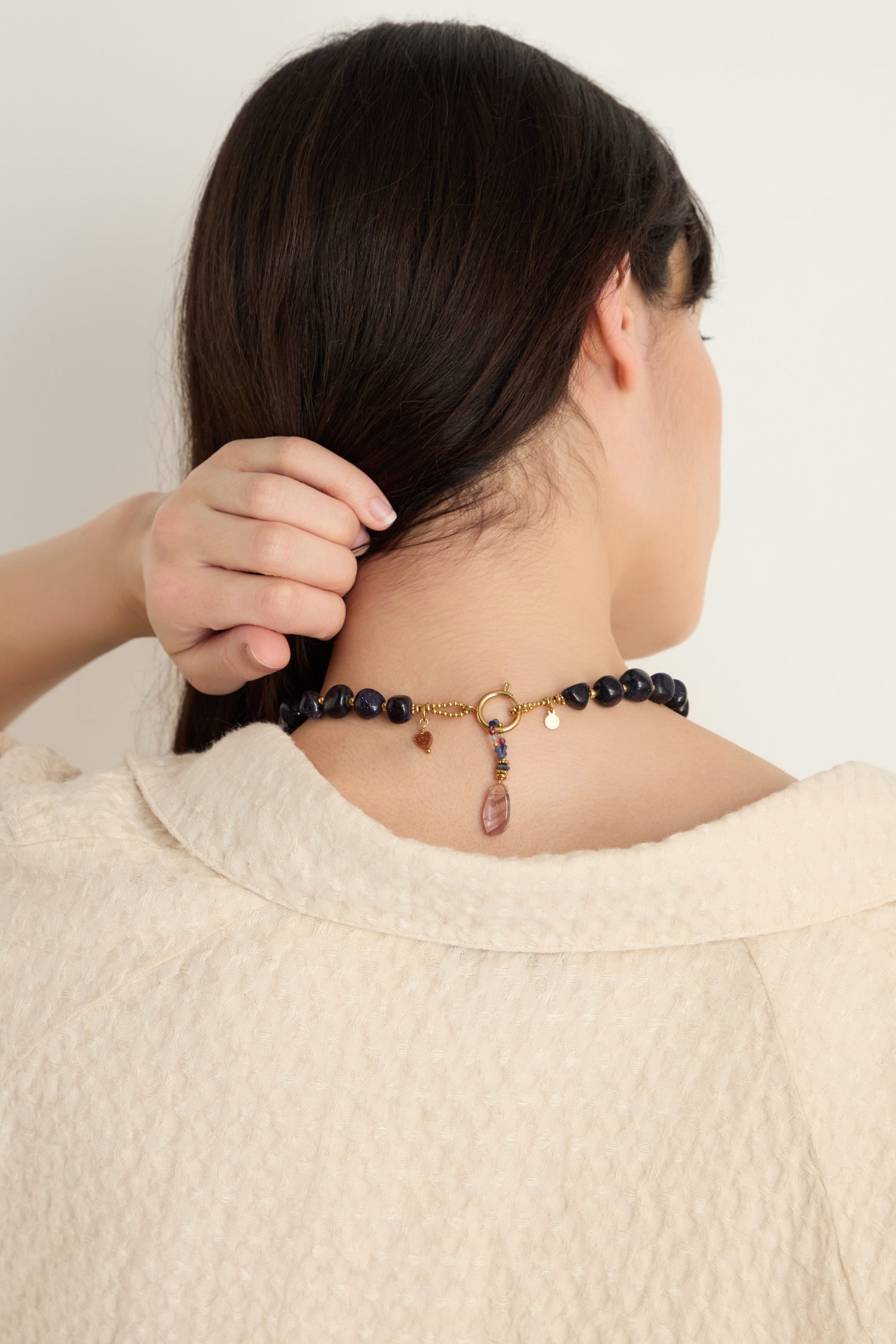 A woman with dark hair, seen from behind, lifts her hair to reveal the Sunstone Bead Necklace featuring semi-precious charms and a gold clasp. She wears a textured cream garment.