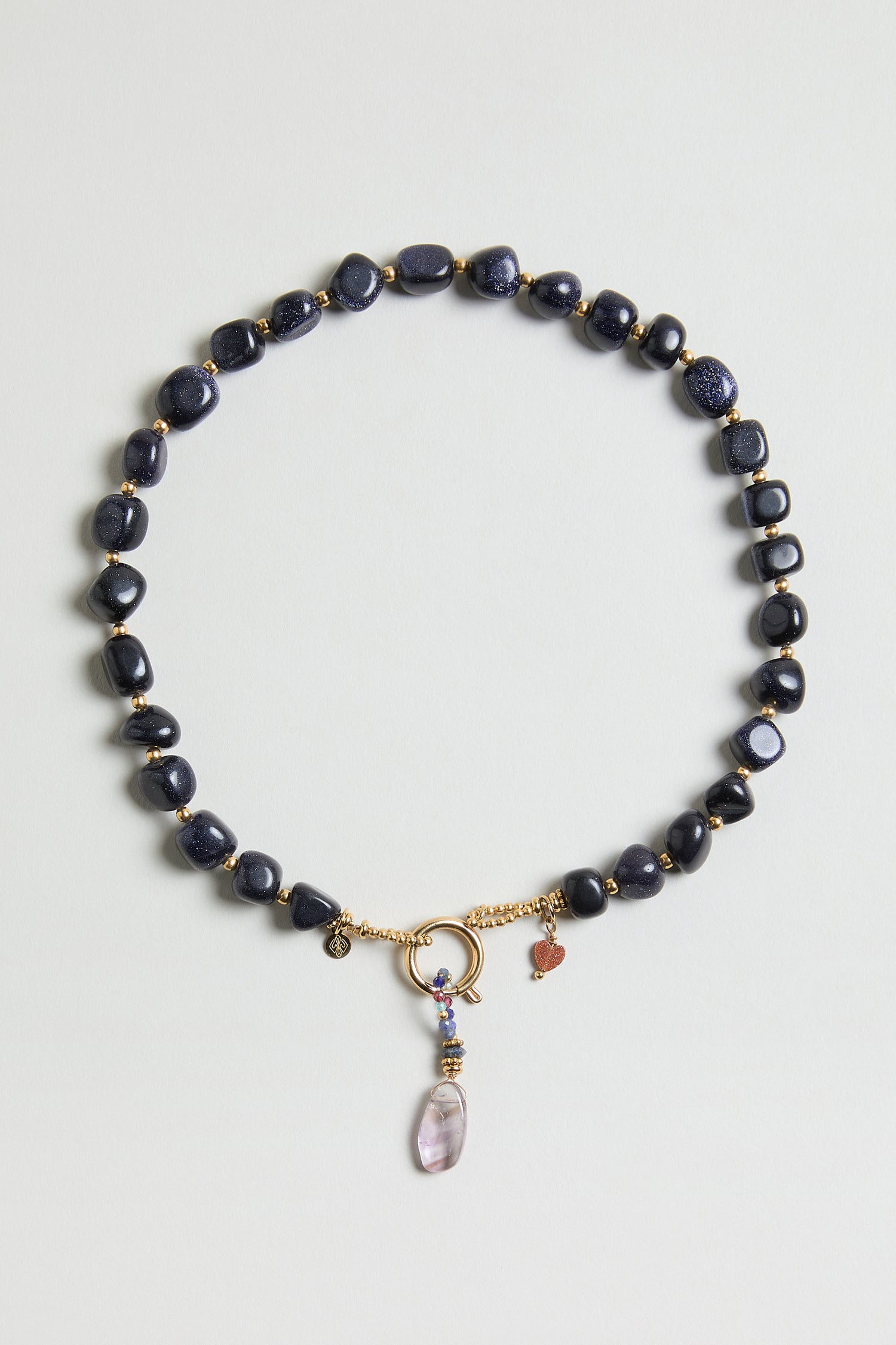 The Sunstone Bead Necklace (MJU33026MG47) features dark blue oval beads, gold accents, a circular gold clasp, small gold and heart charms, and a purple gemstone pendant.