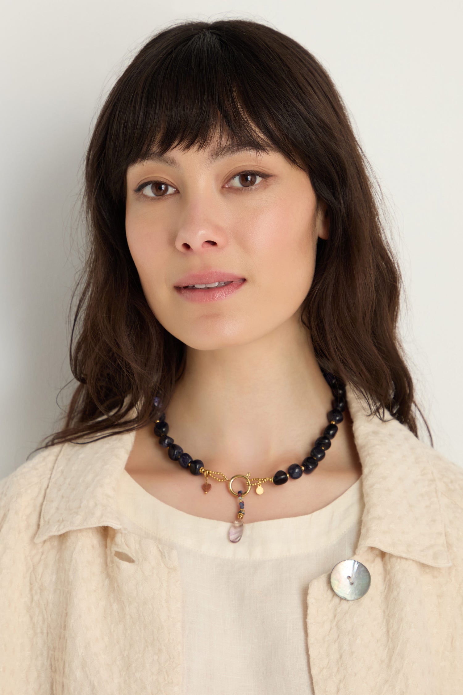 A woman with long dark hair wears a cream textured jacket, a light blouse, a round brooch, and the Sunstone Bead Necklace featuring gold-plated stainless steel and semi-precious charms.