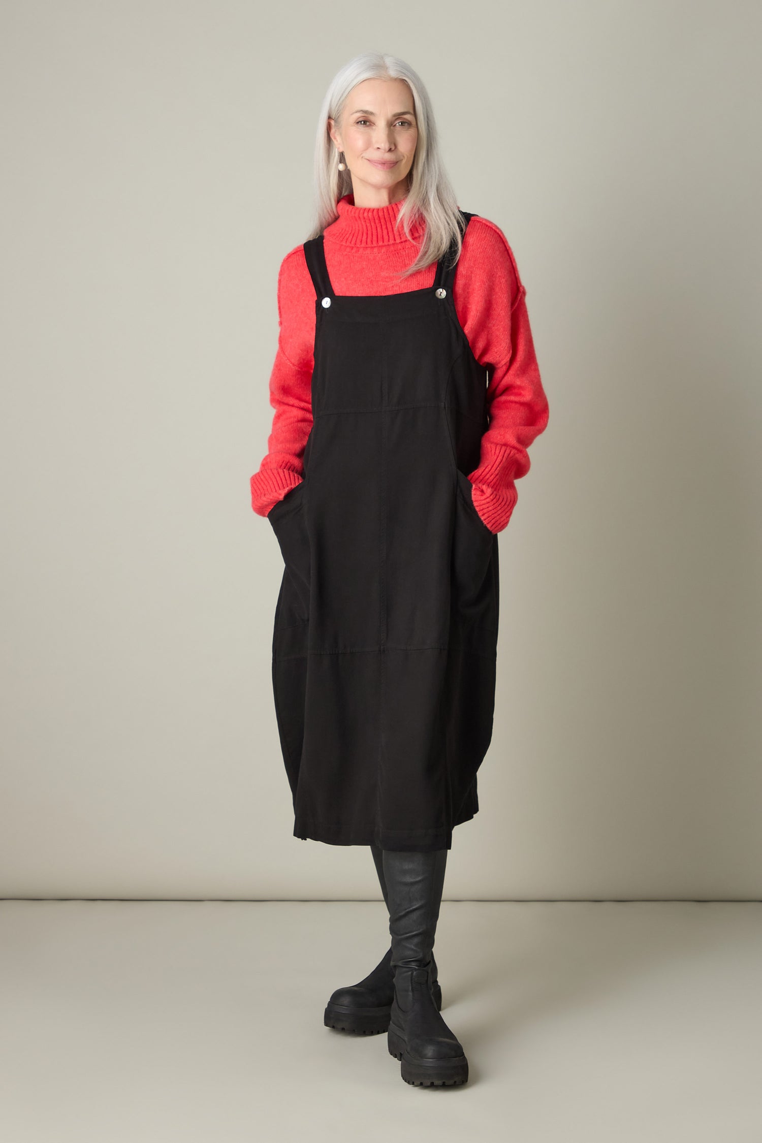A person with long gray hair exudes bohemian charm in a red sweater and a black dress, hands casually tucked in pockets against a plain background.