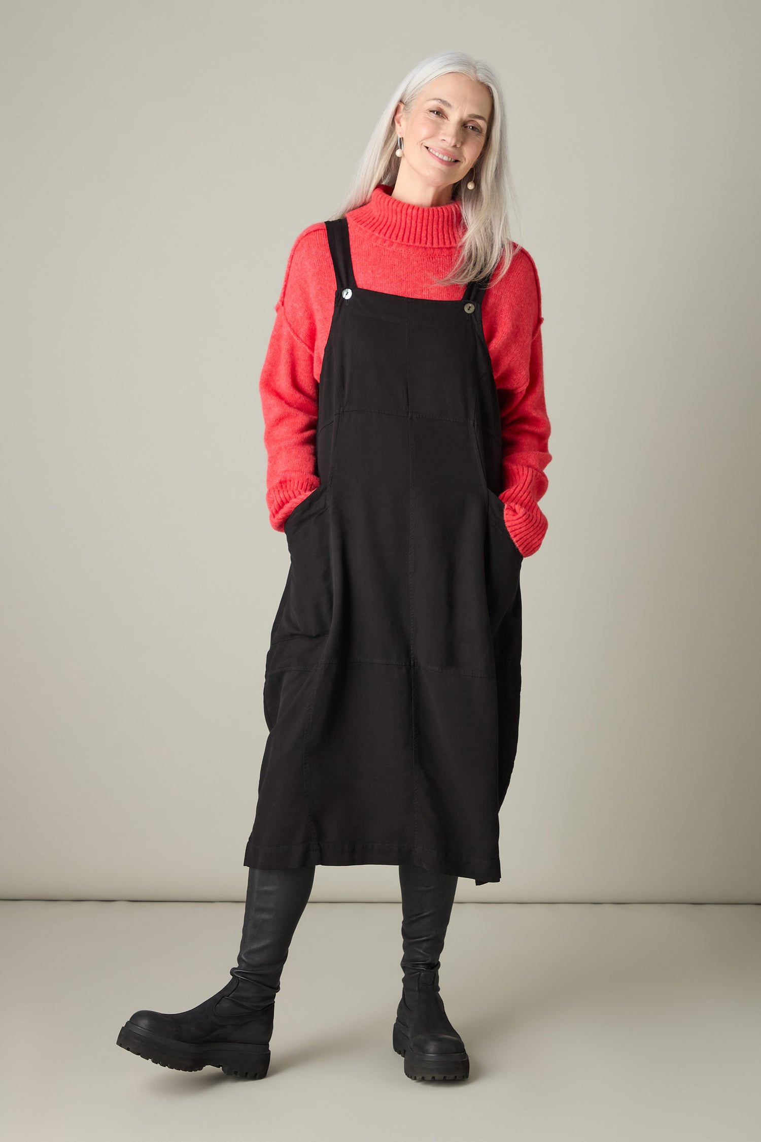 A person with long gray hair exudes bohemian charm in a red sweater, the Fluid Twill Pinafore Dress, black leggings, and boots, standing against a plain background.