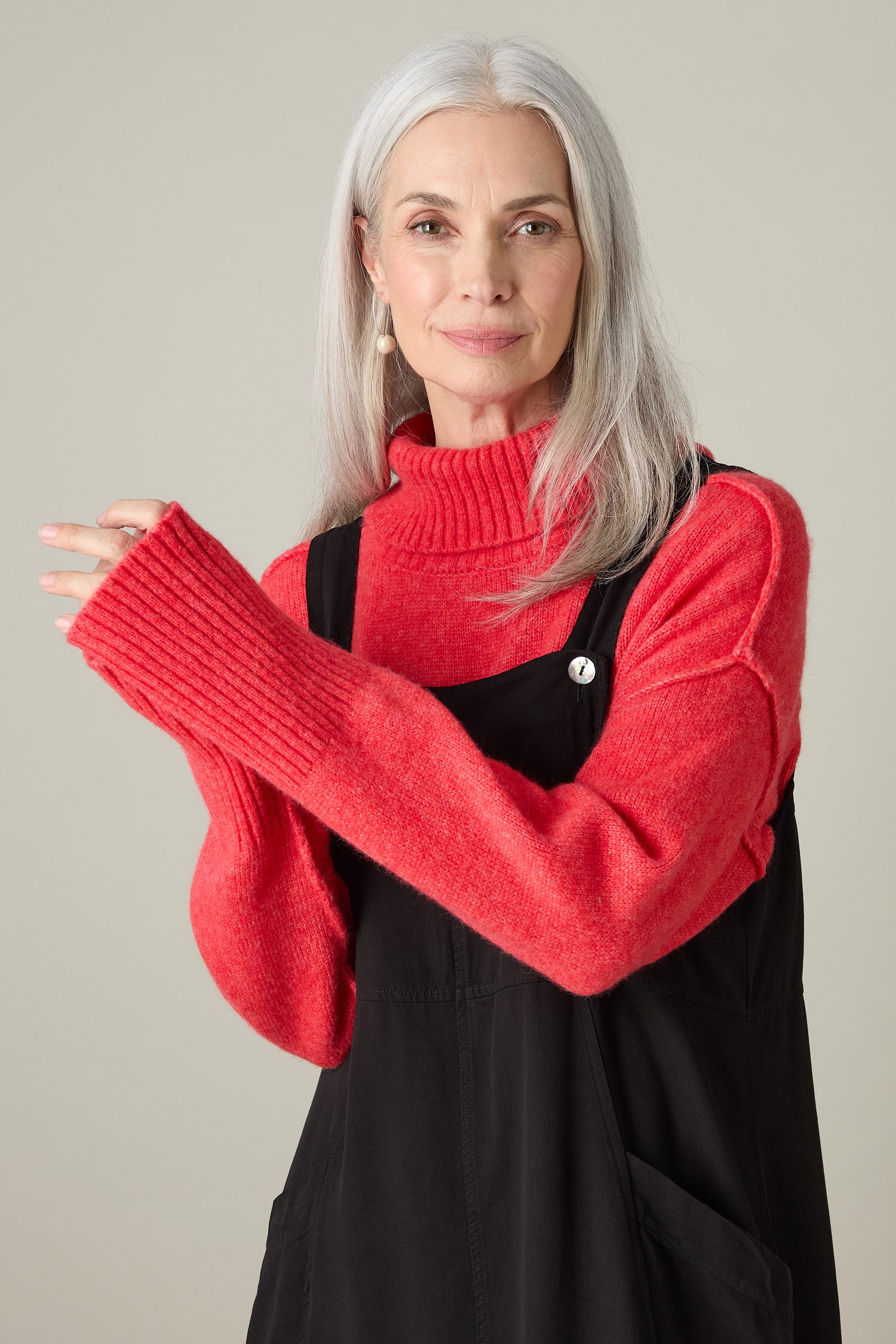 A woman with gray hair, dressed in a red sweater and the Fluid Twill Pinafore Dress, exuding bohemian charm, smiles with her hands gracefully crossed.