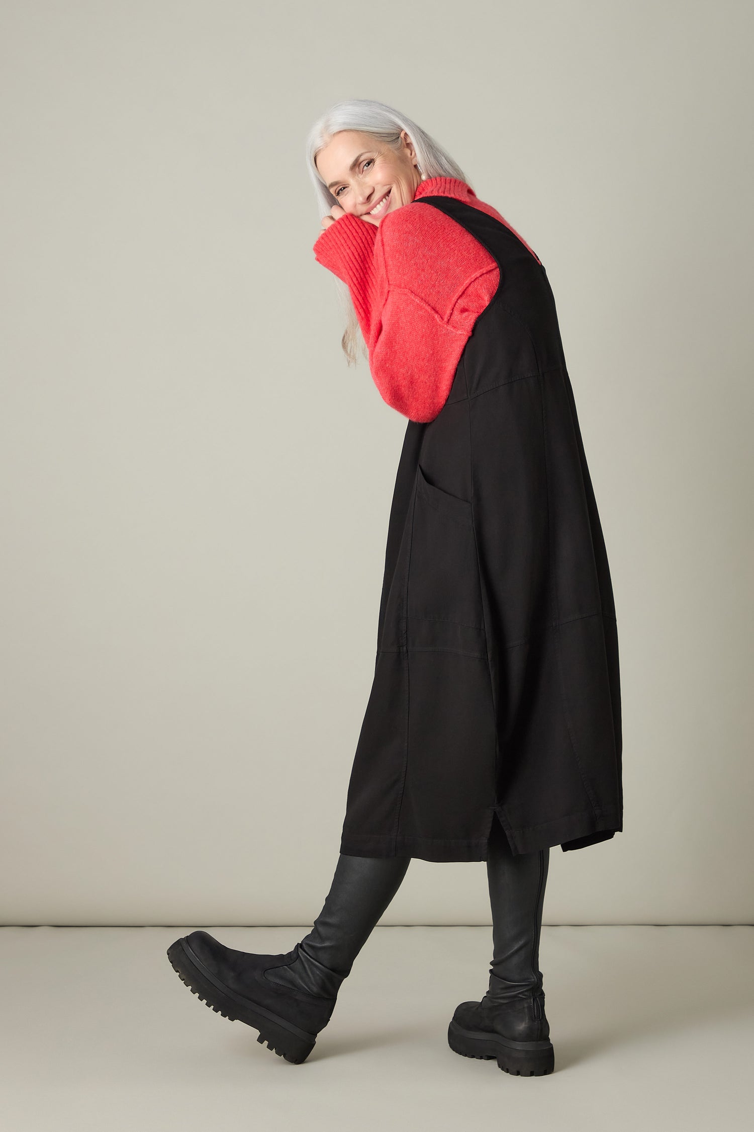 Against a plain backdrop, an individual with long white hair and a red sweater exudes bohemian charm while posing sideways with a smile, styled in the Fluid Twill Pinafore Dress, complemented by black leggings and boots.