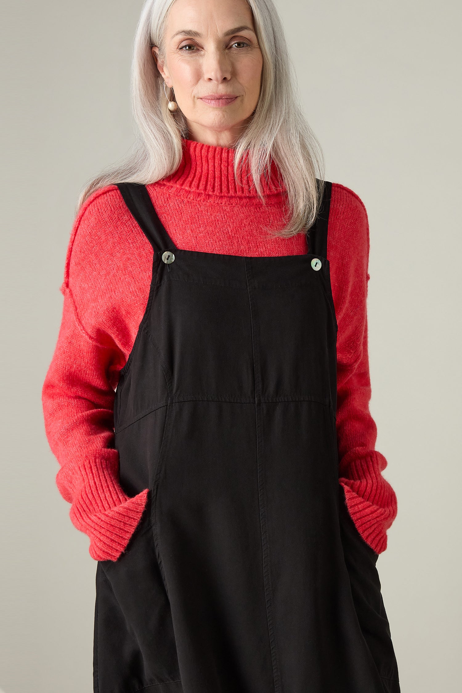 A person with long gray hair, radiating a bohemian charm, stands against a neutral background wearing a red sweater paired with the stylish black Fluid Twill Pinafore Dress.