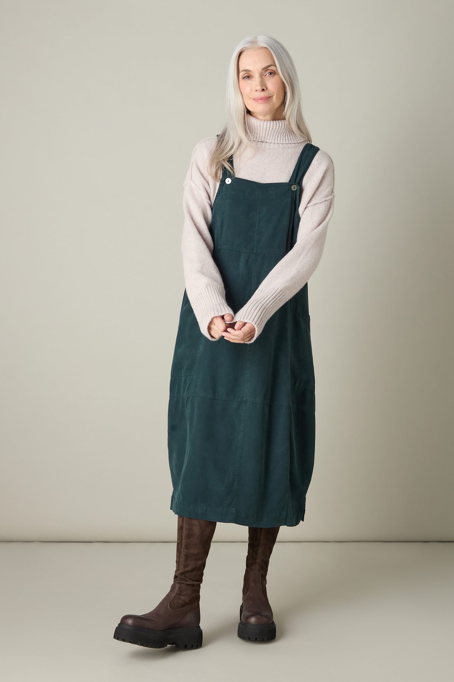 A person with long gray hair stands against a plain background, exuding bohemian charm in the green Fluid Twill Pinafore Dress, paired beautifully with a beige TENCEL turtleneck sweater and brown boots.