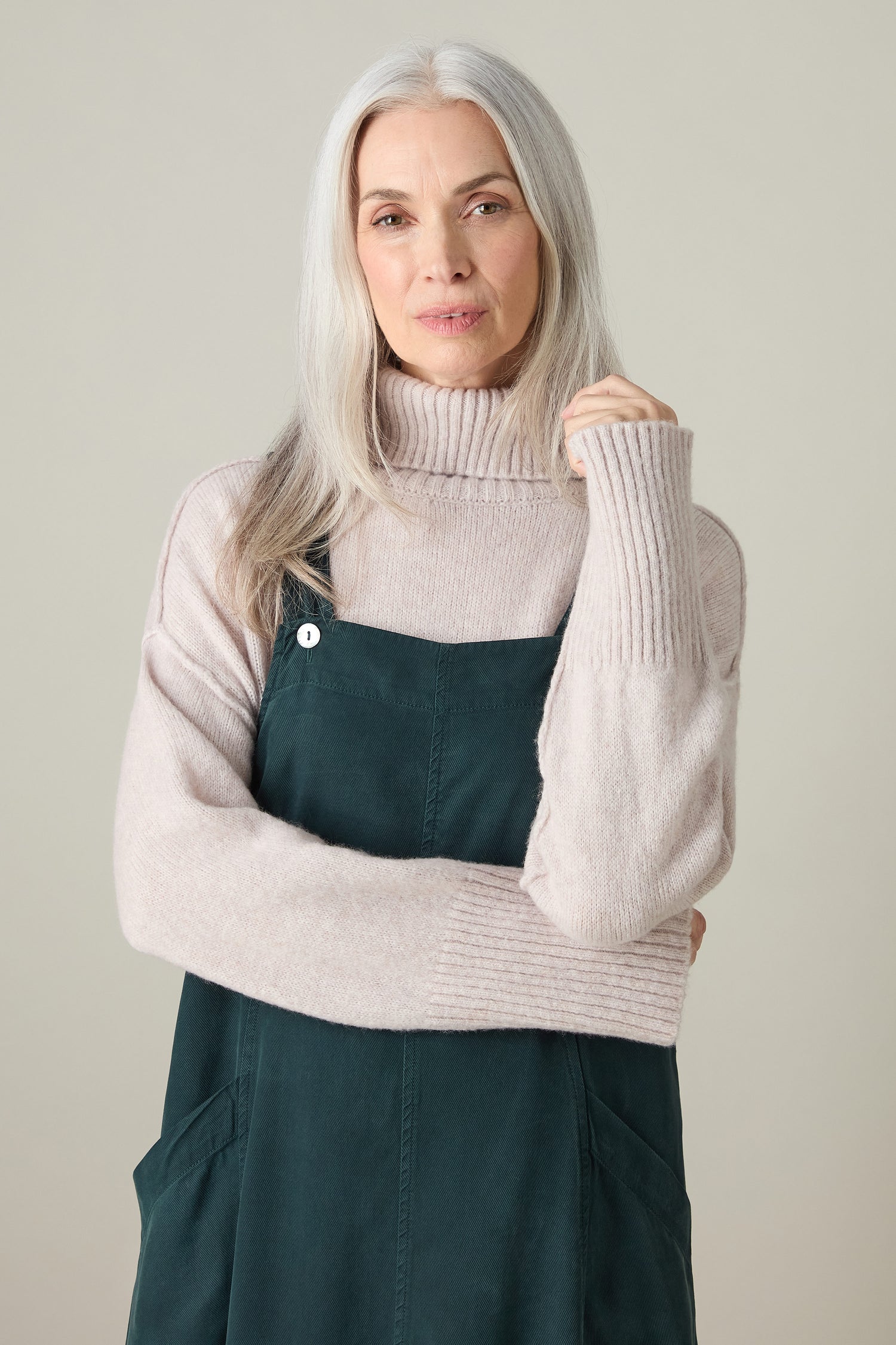 An older woman with long gray hair exudes bohemian charm in a light pink turtleneck and the Fluid Twill Pinafore Dress, standing against a neutral background.