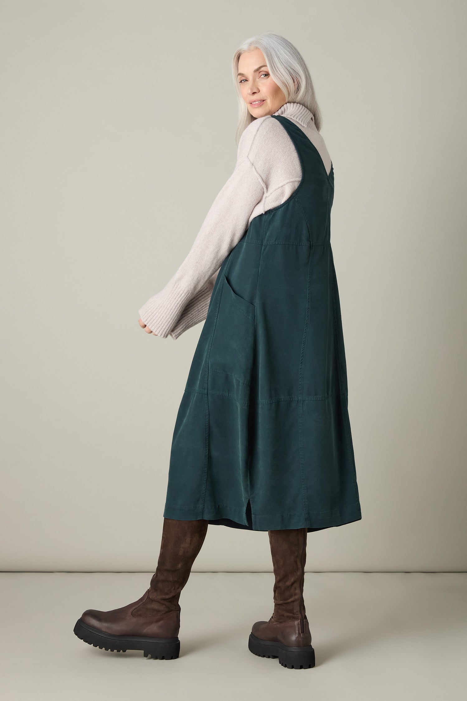 With long gray hair that radiates bohemian charm, a person models the Fluid Twill Pinafore Dress over a beige sweater, paired with tall brown boots, against a simple backdrop.