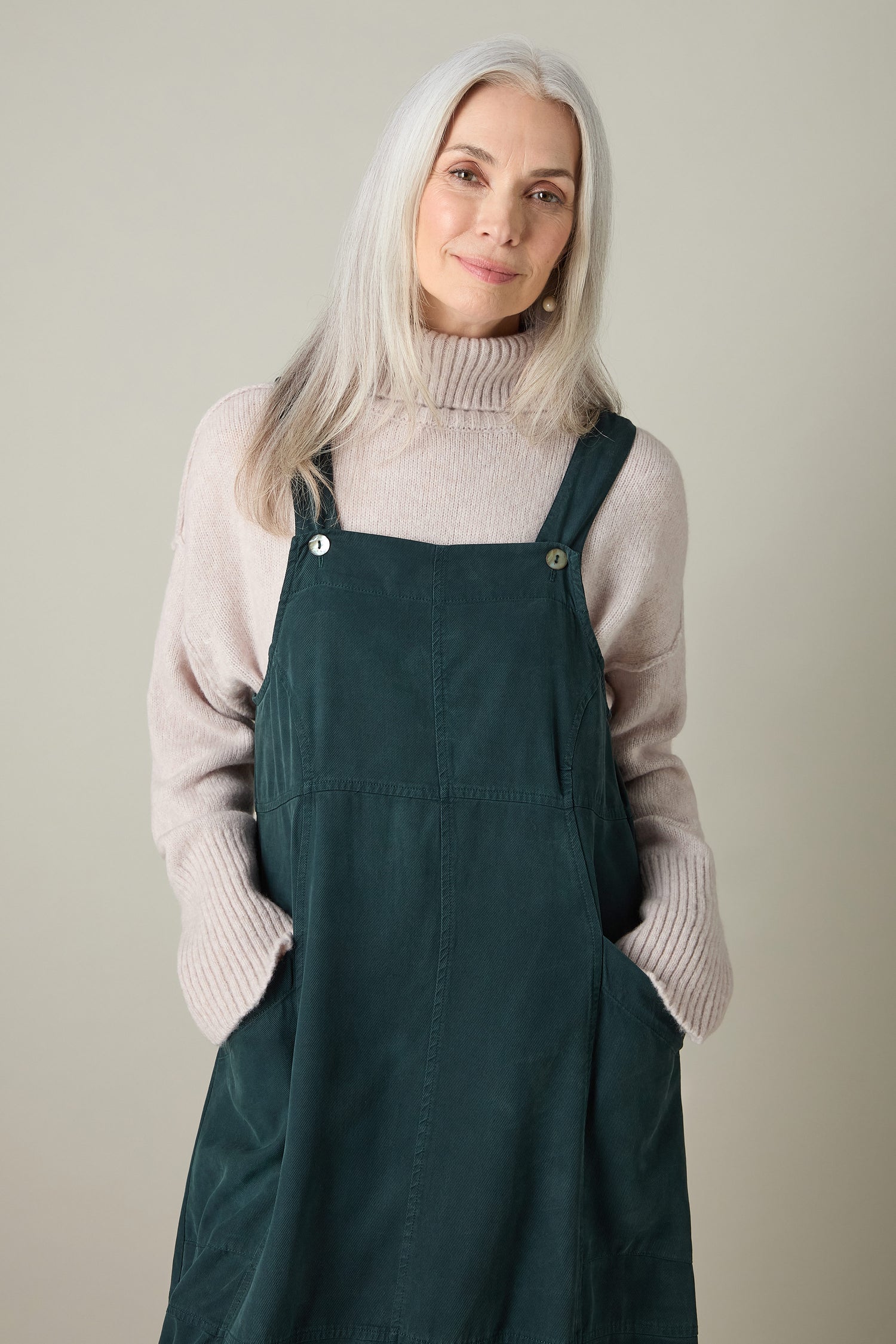 A woman with long gray hair exudes bohemian charm wearing a beige sweater layered under a green Fluid Twill Pinafore Dress, set against a neutral background.