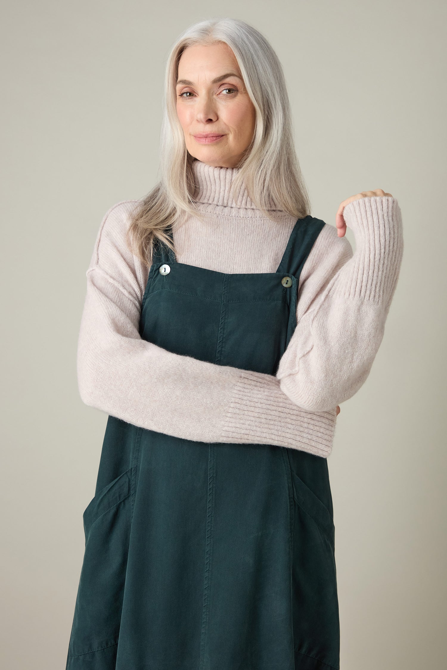 A person with long gray hair exudes bohemian charm in a Fluid Twill Pinafore Dress paired with a light sweater, standing against a neutral background.