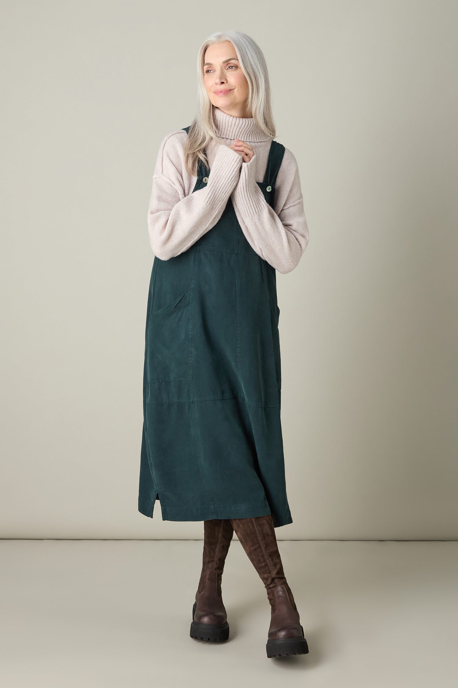 Set against a neutral backdrop, a woman with long gray hair radiates bohemian allure in her beige sweater layered over the green Fluid Twill Pinafore Dress, paired with brown boots.