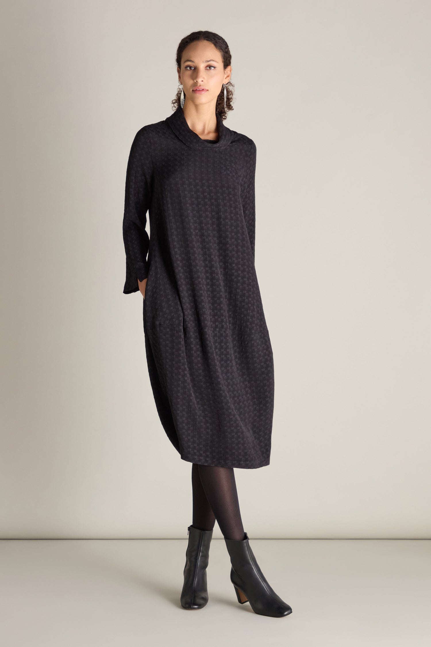 A woman stands against a plain background wearing a long-sleeved, black, textured Small Square Jacquard Bubble Dress with her hands in the pockets and black heeled ankle boots. The bubble silhouette of the dress adds a modern touch to her ensemble.