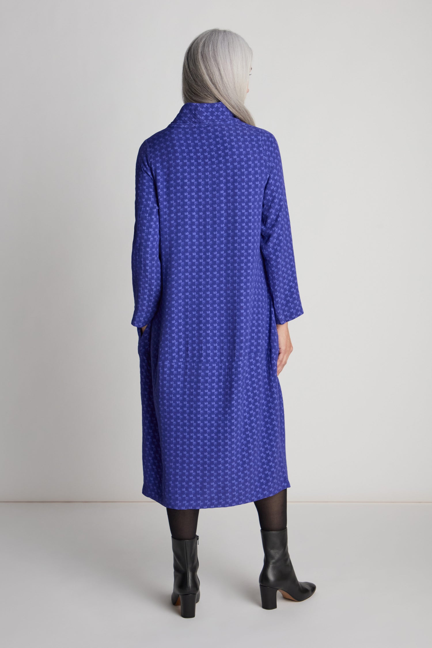 A person with long, gray hair in a Small Square Jacquard Bubble Dress stands facing away from the camera, wearing black tights and black heeled boots.