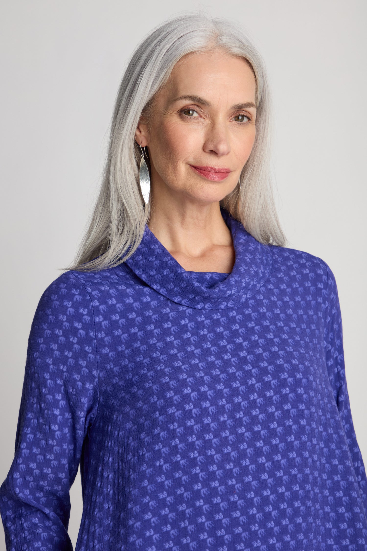 A woman with long gray hair wears a blue patterned blouse and silver earrings, looking directly at the camera against a plain background, reminiscent of a Small Square Jacquard Bubble Dress.