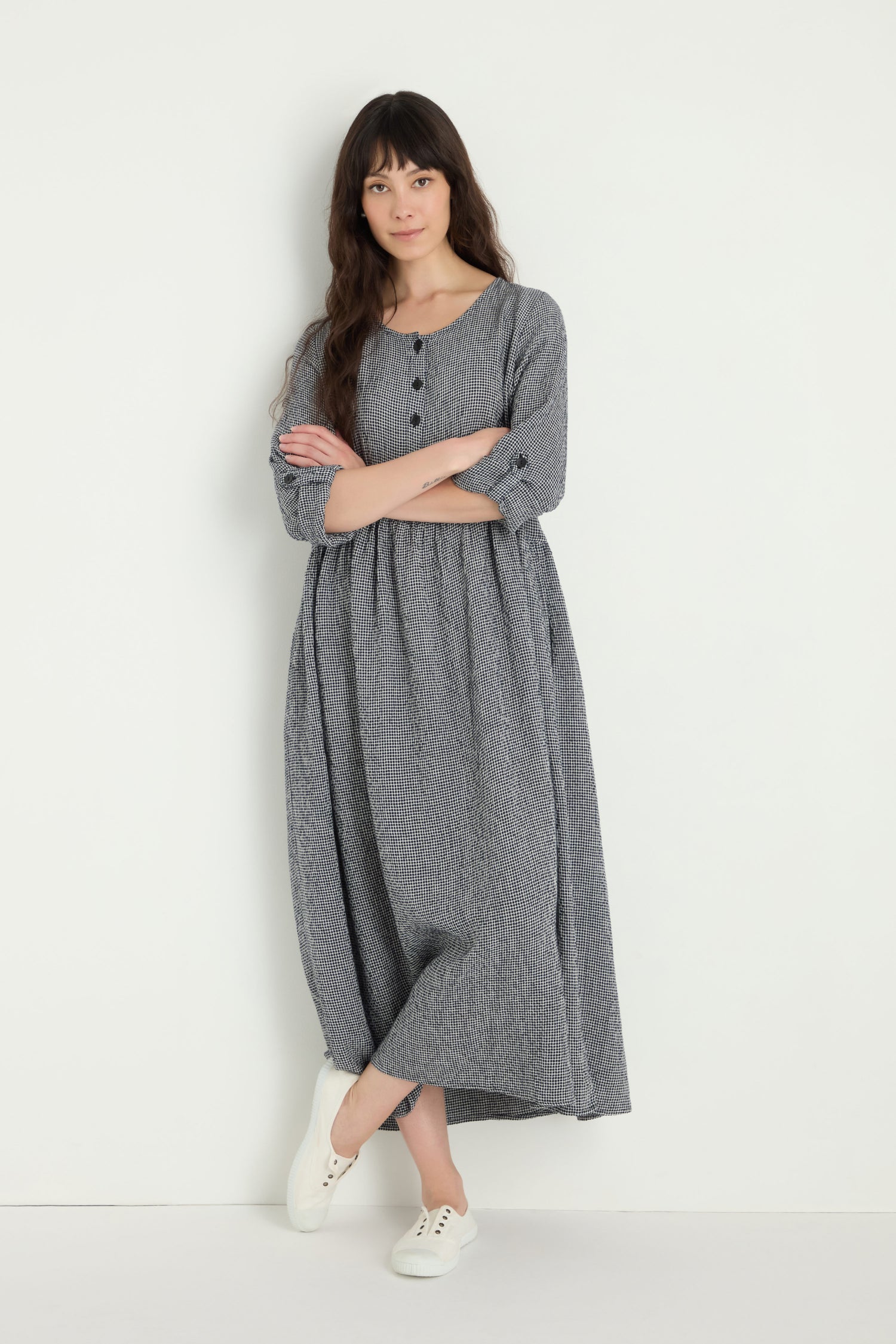 A woman with long dark hair stands against a white wall, arms crossed, wearing the Micro Gingham Bubble Dress in gray with buttons and white sneakers.