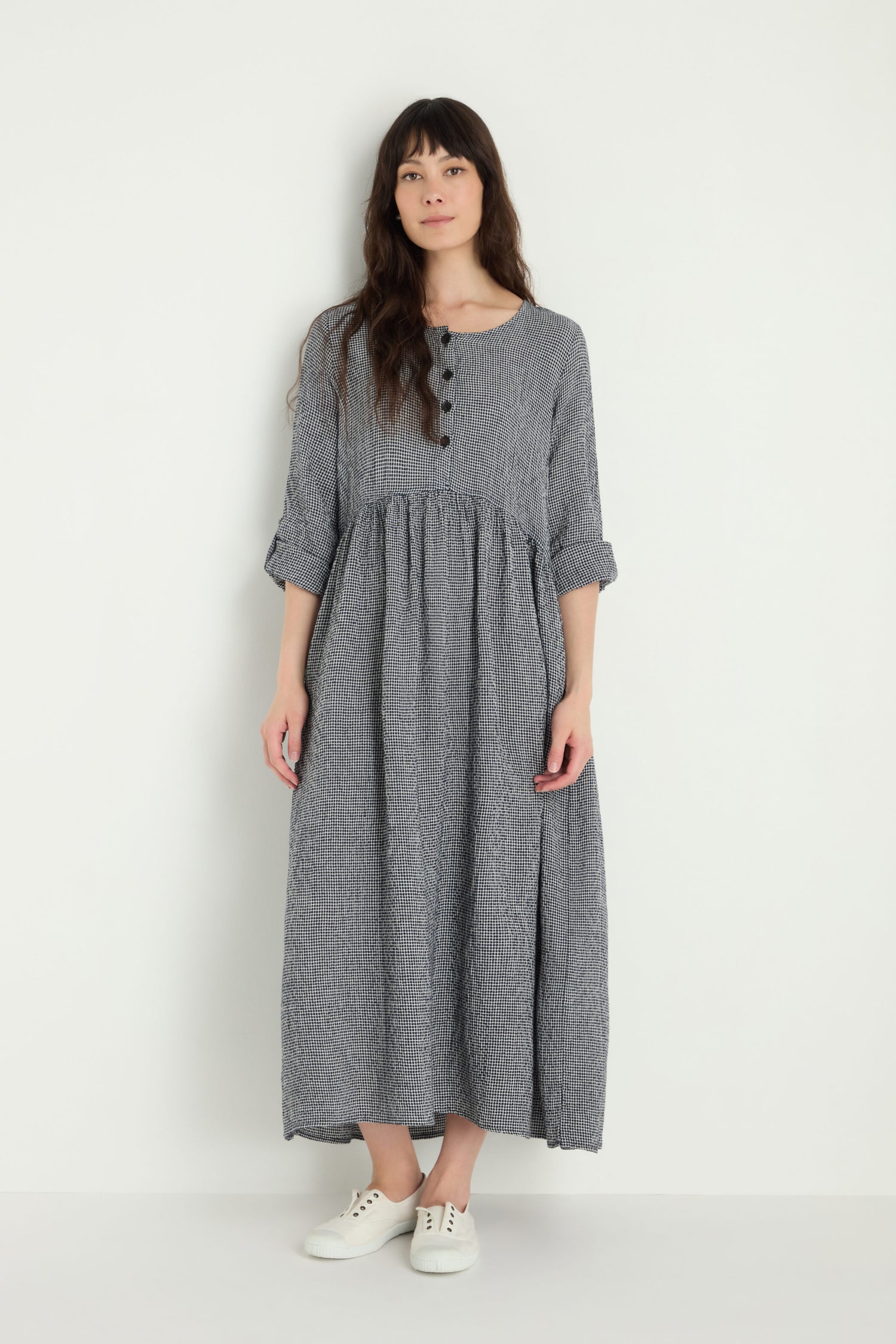 A woman stands against a plain white background, wearing the Micro Gingham Bubble Dress—an ankle-length, long-sleeved gray dress with buttons—paired with white sneakers.