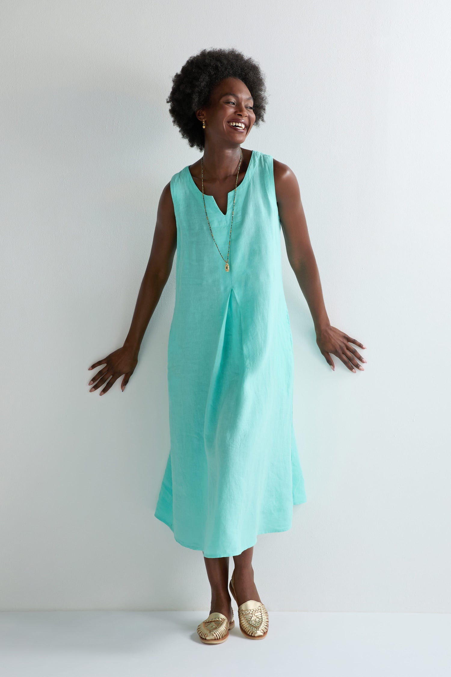 Smiling woman stands by a white wall in the Linen Notch Neck Sleeveless Dress—an aqua A-line silhouette—styled with a gold necklace and woven gold slip-on shoes, making it perfect for your summer wardrobe.