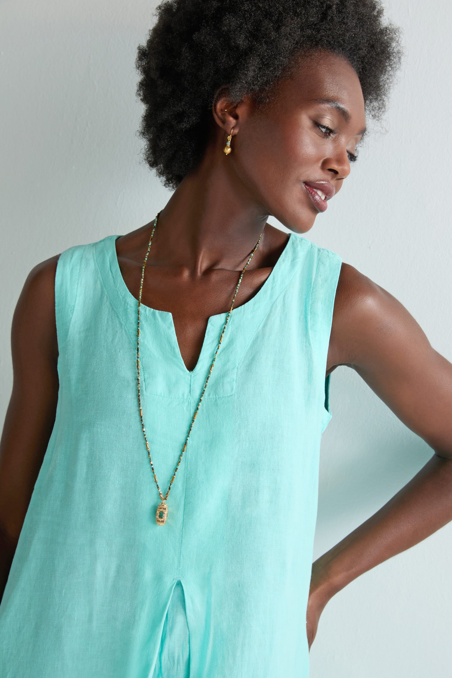 A woman in a Linen Notch Neck Sleeveless Dress, styled with a long beaded necklace and gold earrings, stands against a pale background, her summery look completed by a gentle smile as she gazes to the side.