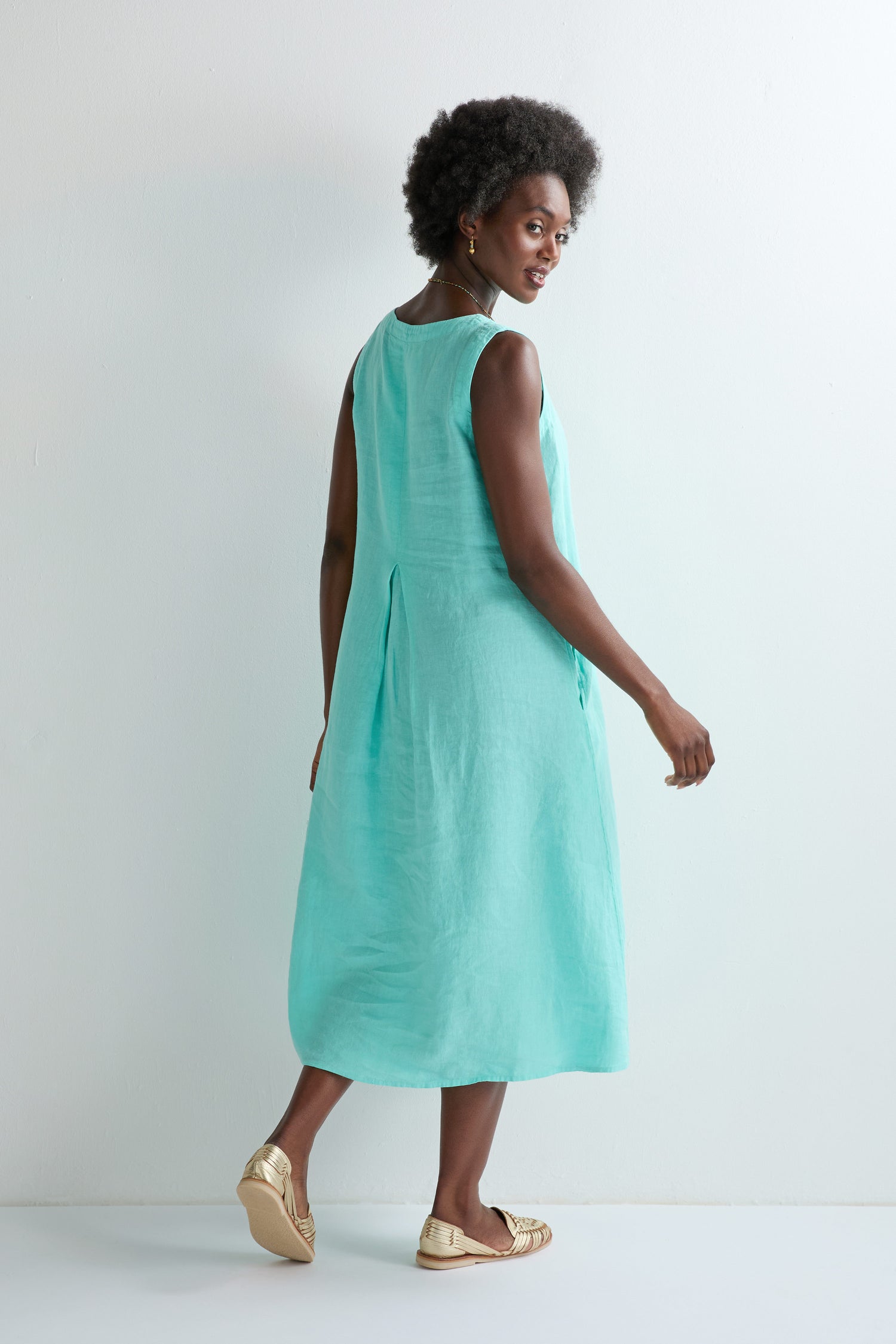 A woman stands sideways, looking back, wearing the Linen Notch Neck Sleeveless Dress in light turquoise with an A-line silhouette and gold slip-on shoes—a chic summer wardrobe choice against a plain light background.