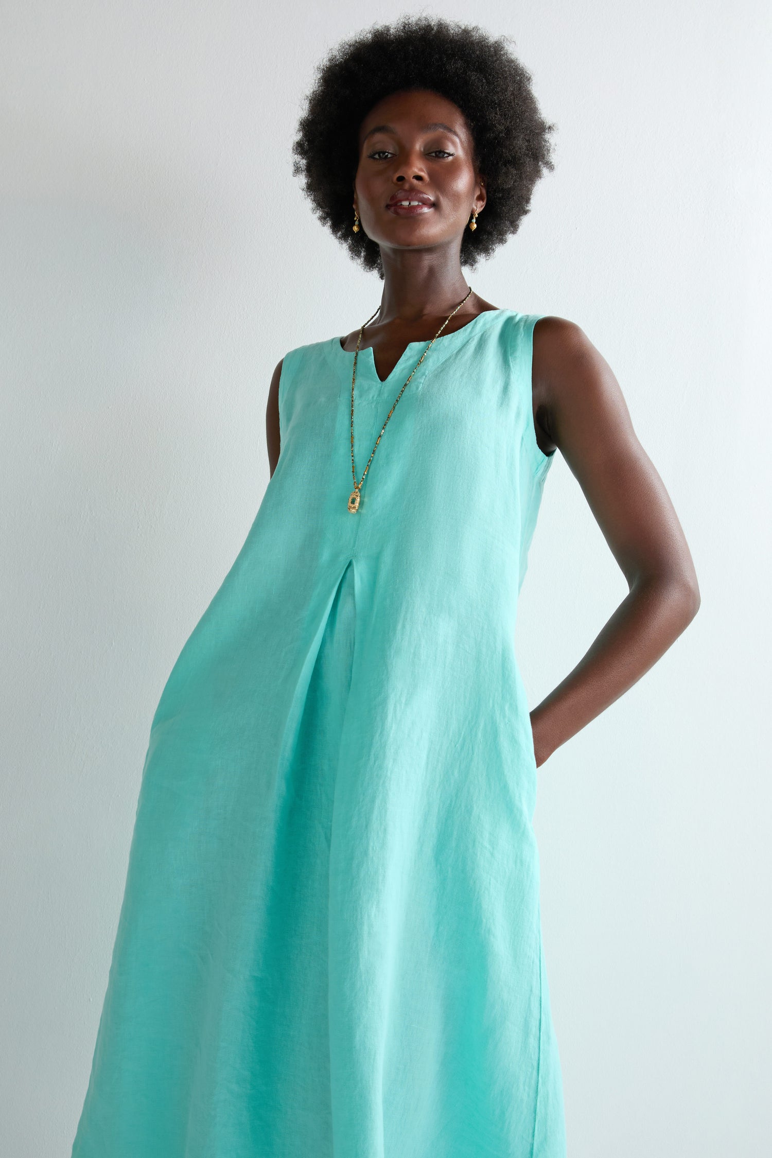 A person stands against a light background wearing the Linen Notch Neck Sleeveless Dress in light blue, featuring side pockets and an A-line silhouette—an effortless summer wardrobe staple, paired with a necklace.
